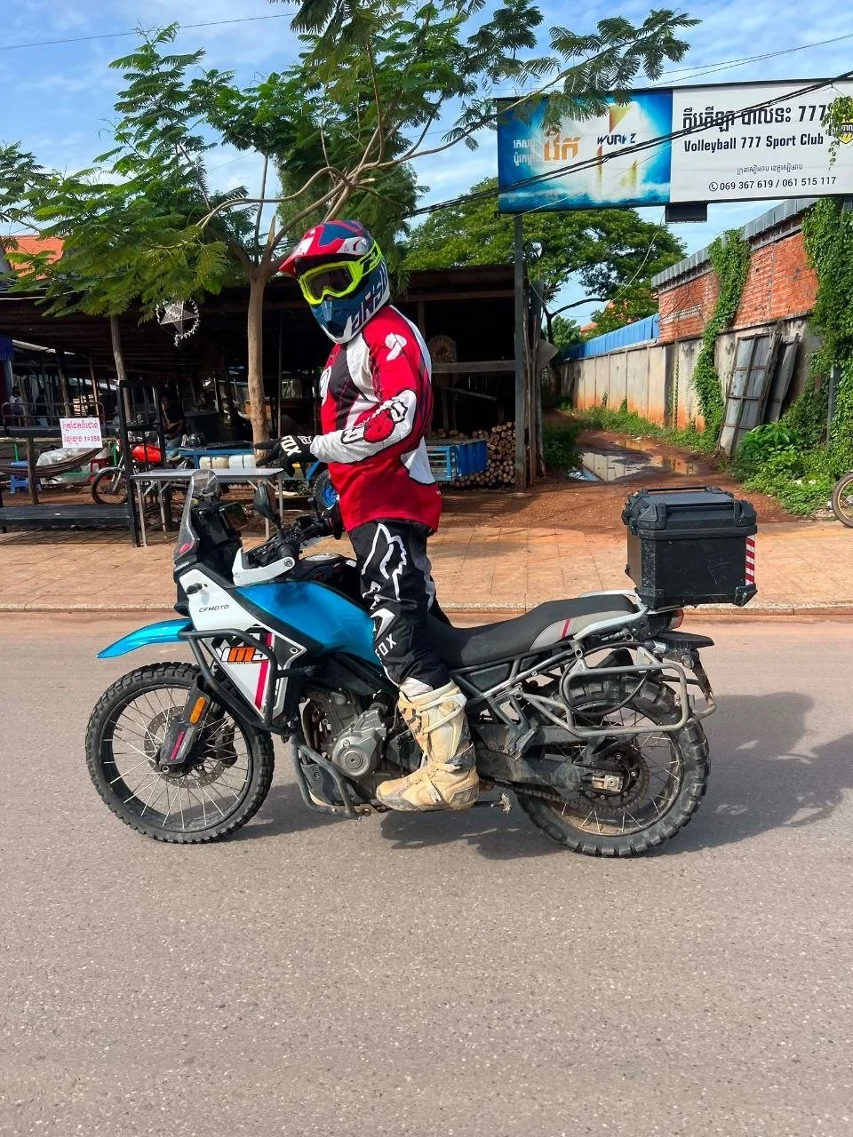 CFMoto MT450 adventure motorcycle with a rider wearing full riding gear standing and looking towards the camera on a street in Siem Reap, Cambodia.