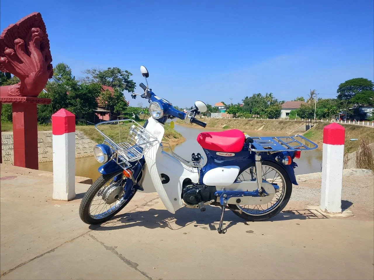 Honda Super Cub at a corner of a bridge over the Siem Reap River with a blue sky.