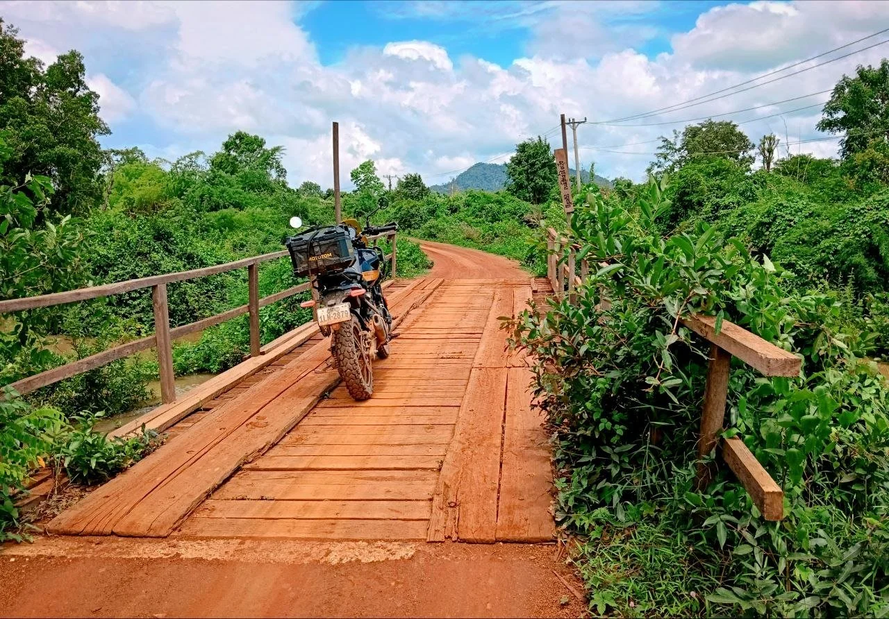 Image of a Royal Enfield Himalayan 450 on a wooden bridge with a mountain view in a green natural Cambodian landscape with a blue sky.
