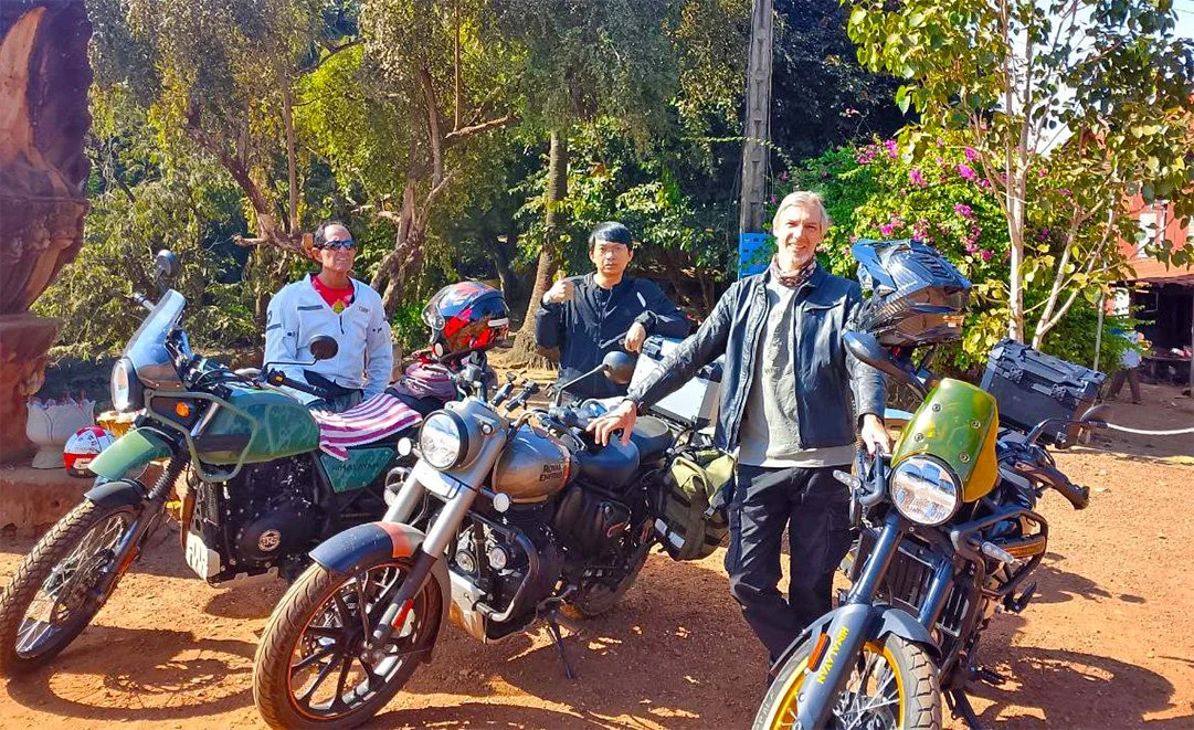 Riders and motorbikes posing in front of the green nature, red dirt and a naga sculpture of Praptos bridge in Cambodia