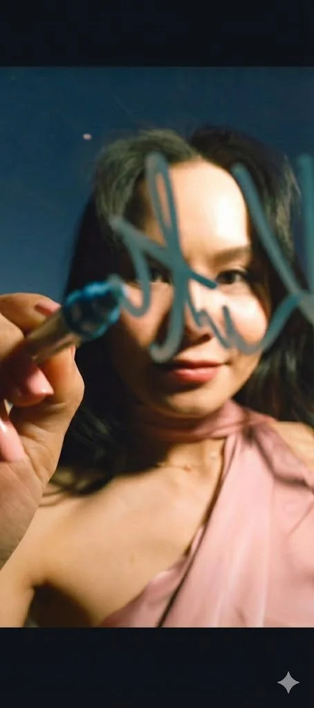 A woman in a white dress joyfully gestures next to a person holding a bouquet of white flowers covering their face, standing against a rustic wooden wall.