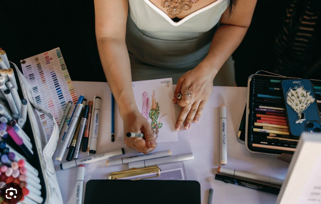 A woman in a white dress joyfully gestures next to a person holding a bouquet of white flowers covering their face, standing against a rustic wooden wall.