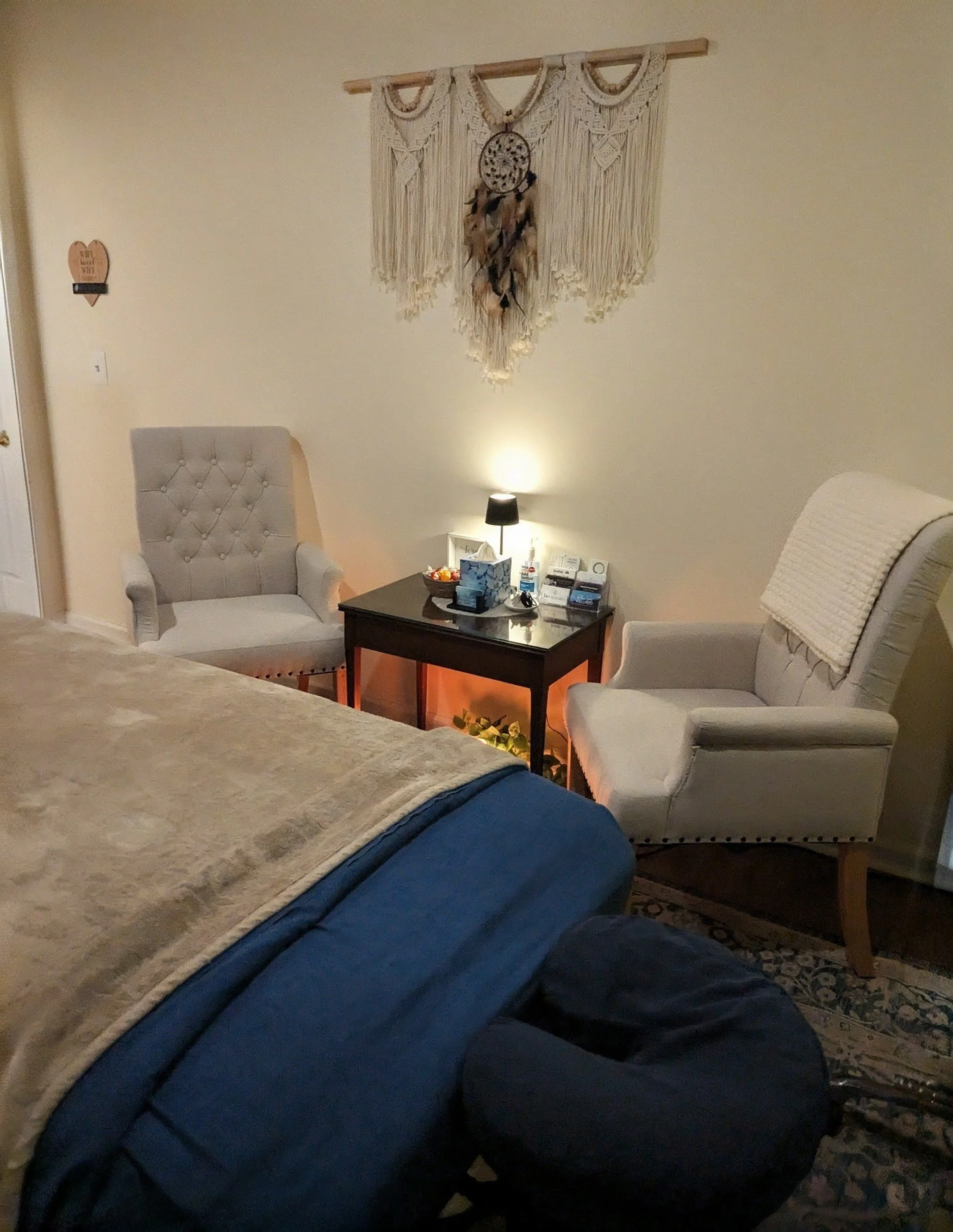 A cozy living room corner with a blue couch, beige armchair, and a dark wood side table with a tissue box, snacks, and a small device. A wall decoration with a dreamcatcher is hanging, and a small lamp provides soft lighting. The room has a cream-colored wall, a patterned rug, and a blanket on the armchair.