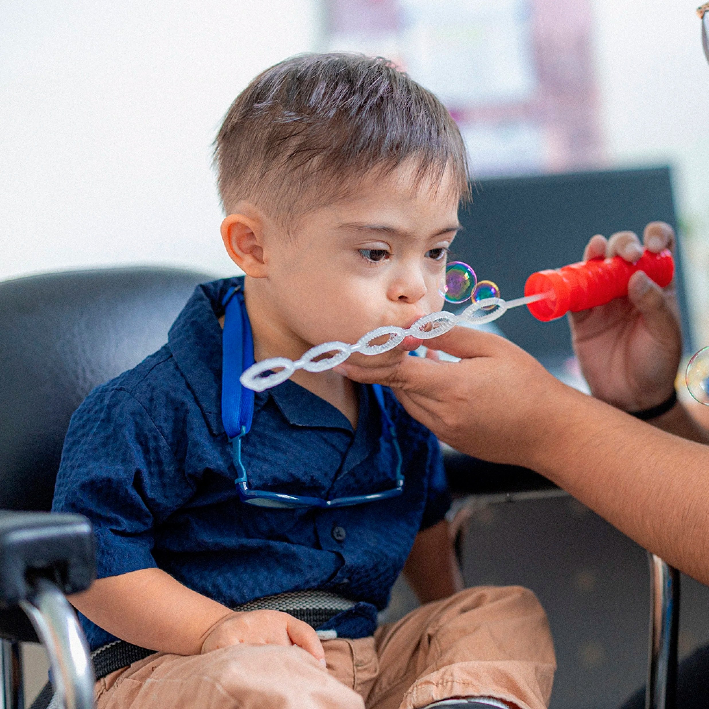 A young boy with short brown hair is sitting in a tall chair , wearing a denim shirt and khaki pants. He is blowing bubbles with a bubble wand, and there are several bubbles floating around his face. The background is blurred,  indoor setting.