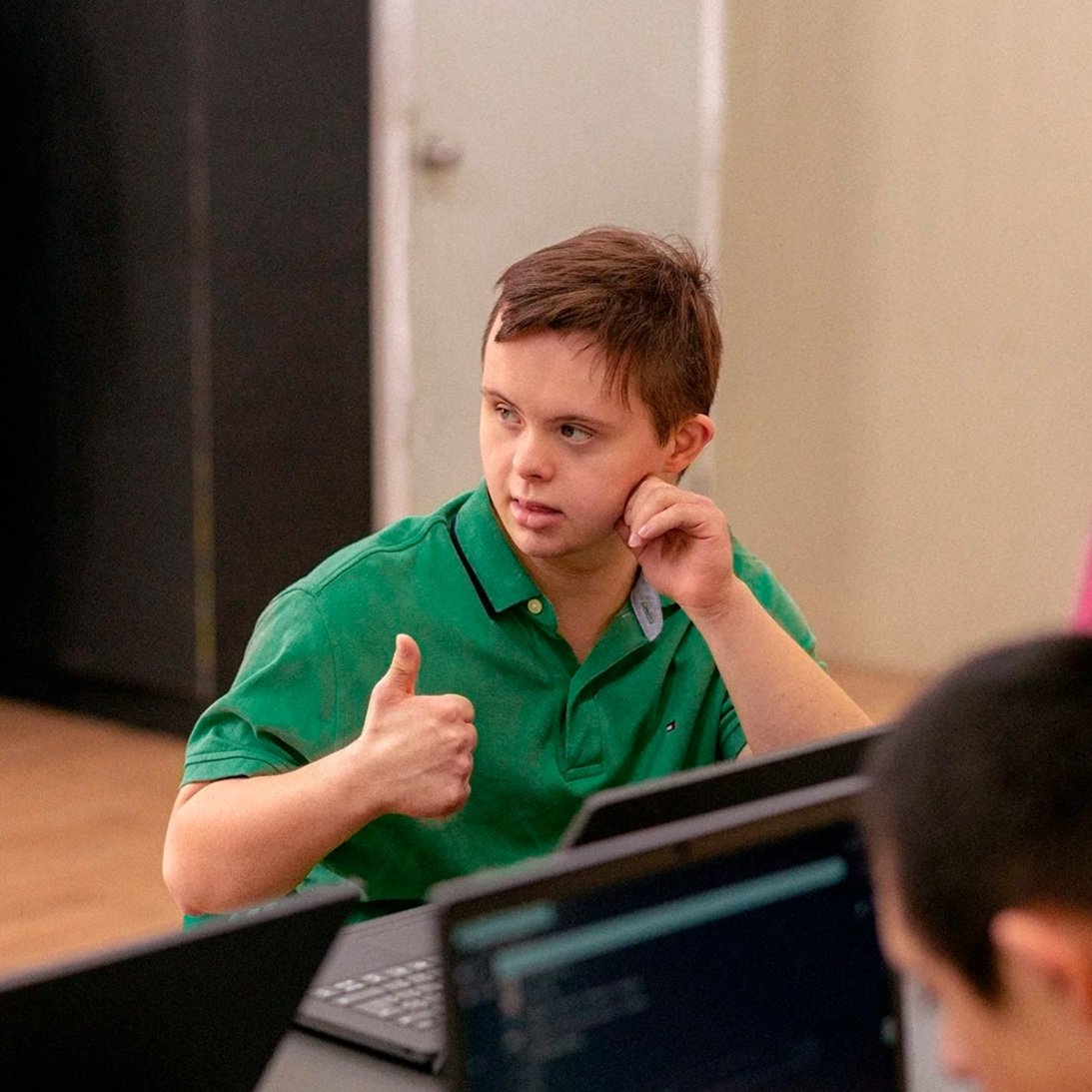 A young boy with short brown hair, wearing a green polo shirt, giving a thumbs-up with his right hand while touching his face with his left hand, sitting in front of a laptop in a classroom . Tedi vanguardia en sindrome de down