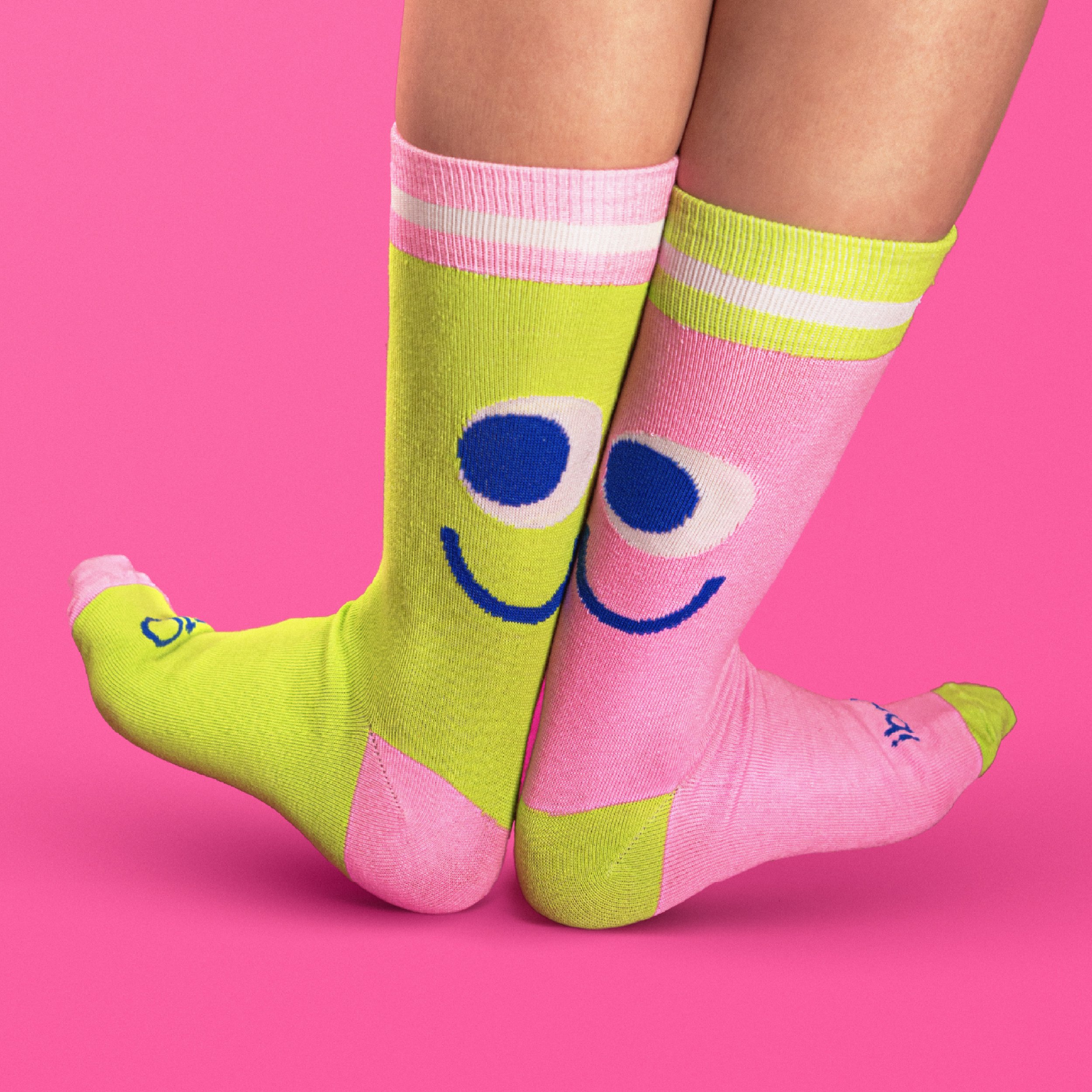 Close-up of legs wearing colorful knee-high socks, one pink with white stripes and the other yellow-green with face cartoon design, against a pink background. Tedi vanguardia en sindrome de down