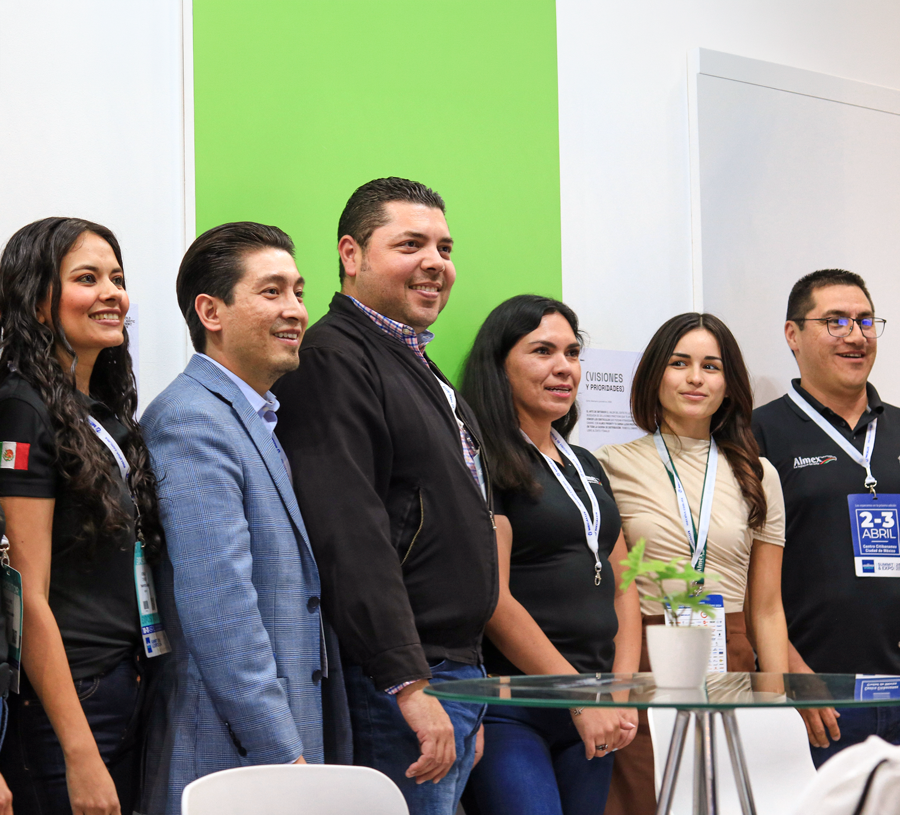 Grupo de seis personas posando para una foto en un evento, detrás de una mesa de vidrio con una planta en maceta blanca, en un fondo de paredes blancas y una pared verde.