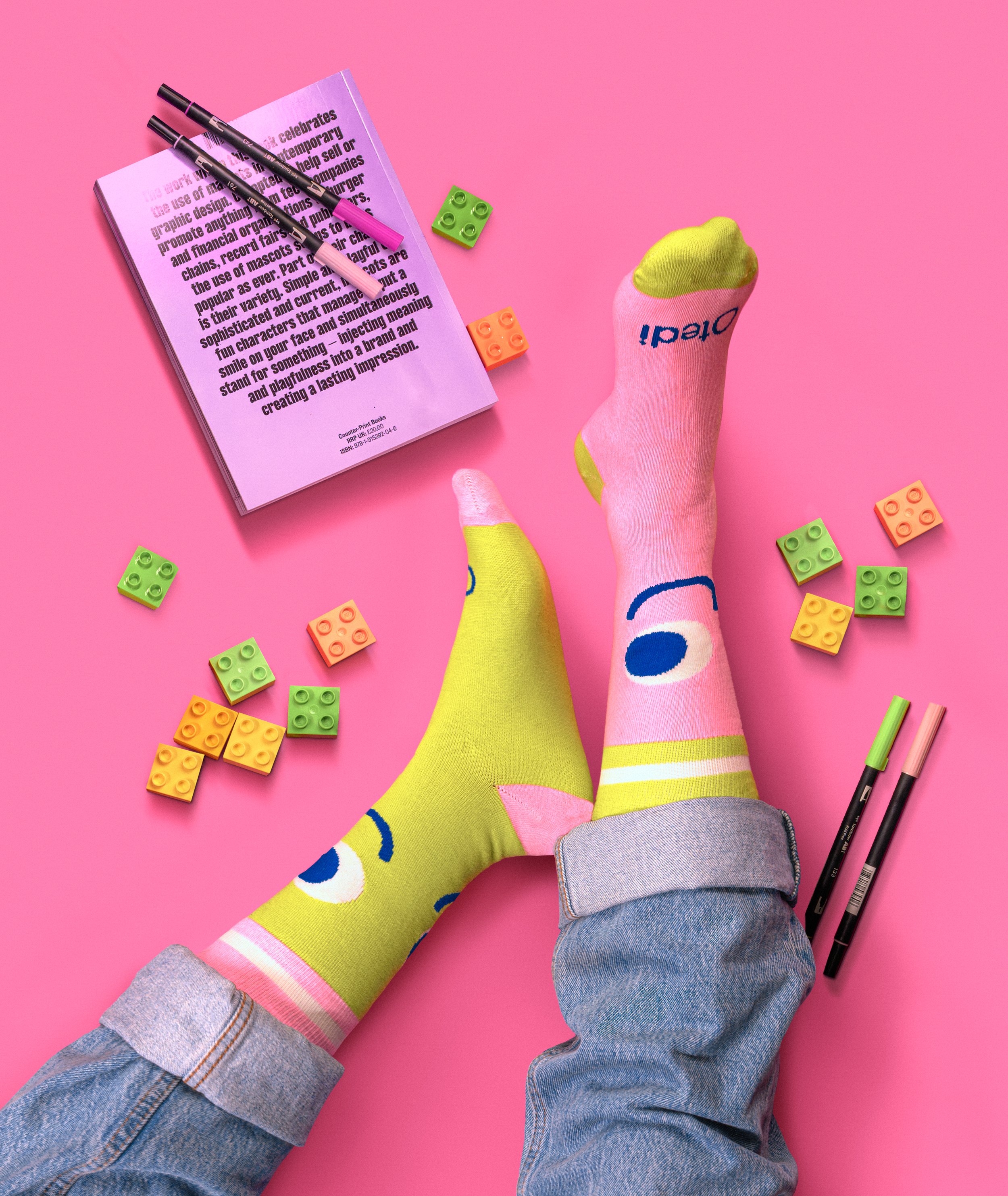 Colorful socks on feet, surrounded by building blocks, markers, a book, on a pink background. Tedi vanguardia en sindrome de down