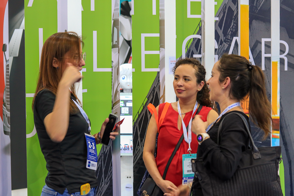 Tres mujeres en una feria o conferencia, conversando y mostrando credenciales, con fondo de carteles y pantallas digitales.