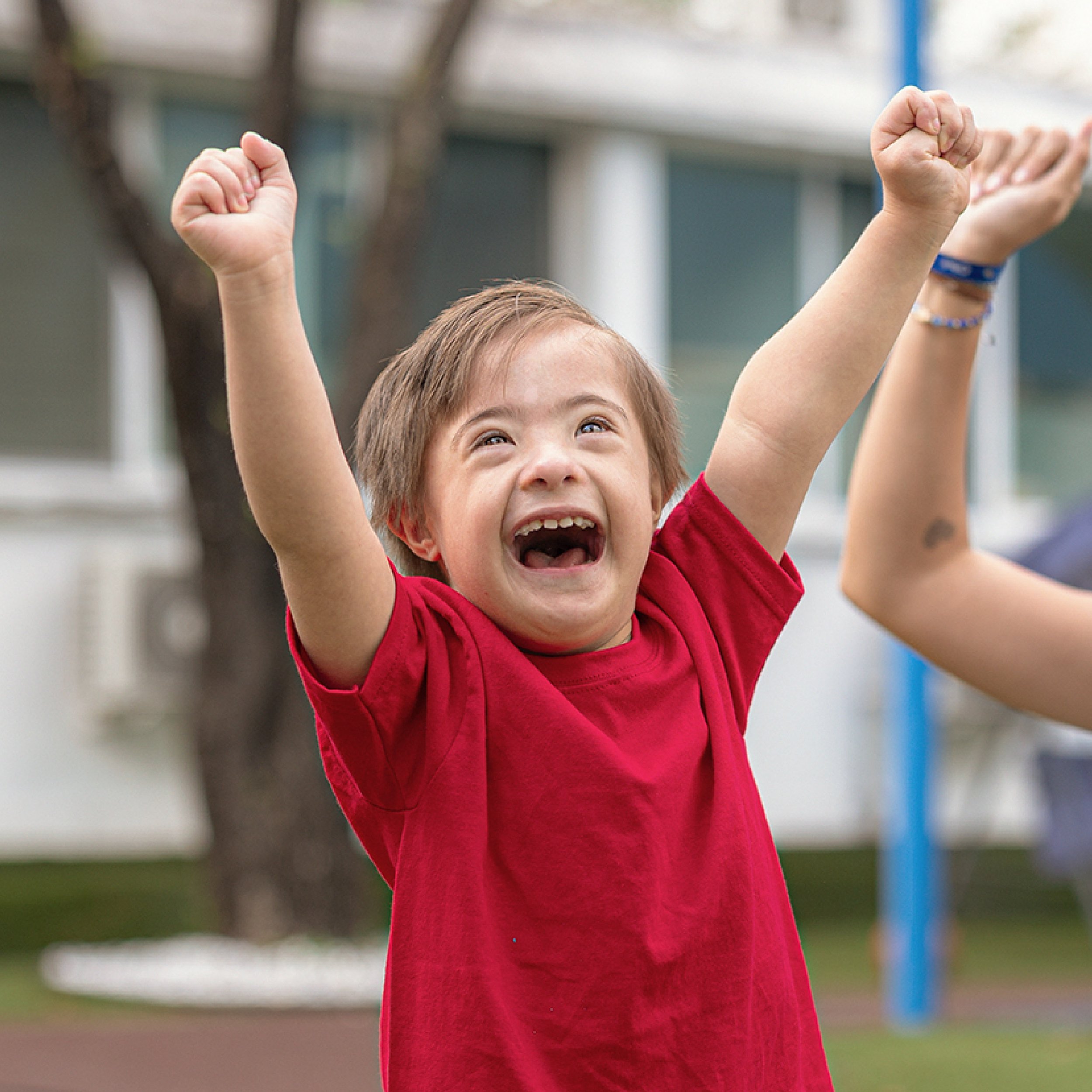 A young boy with light brown hair smiling and raising her arms in the air at a playground. Tedi vanguardia en sindrome de down