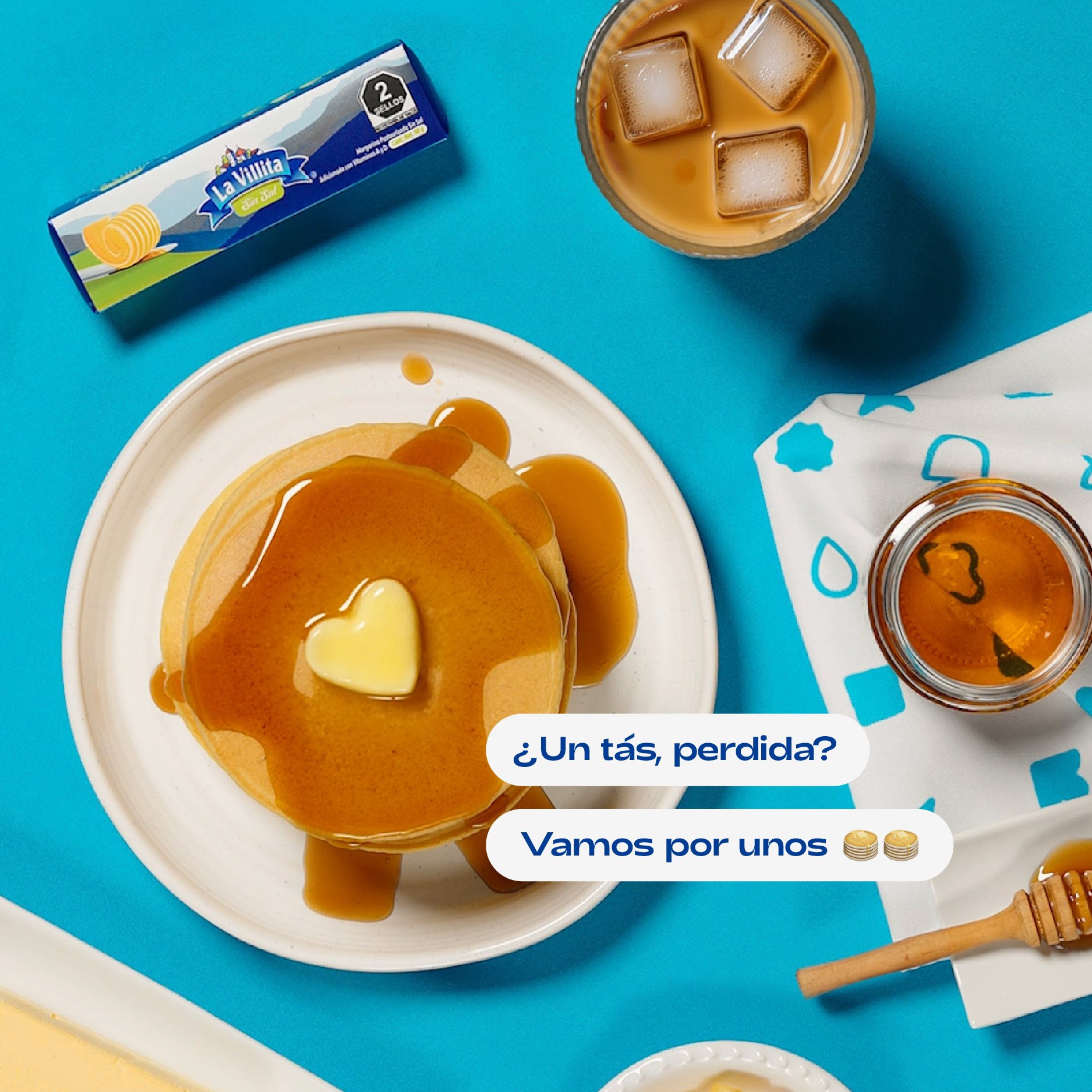 A stack of pancakes with caramel syrup and a heart-shaped dollop of butter on top, on a white plate. Next to it, a glass of iced coffee with ice cubes. The setup is on a bright blue surface with a blue and white patterned cloth nearby.