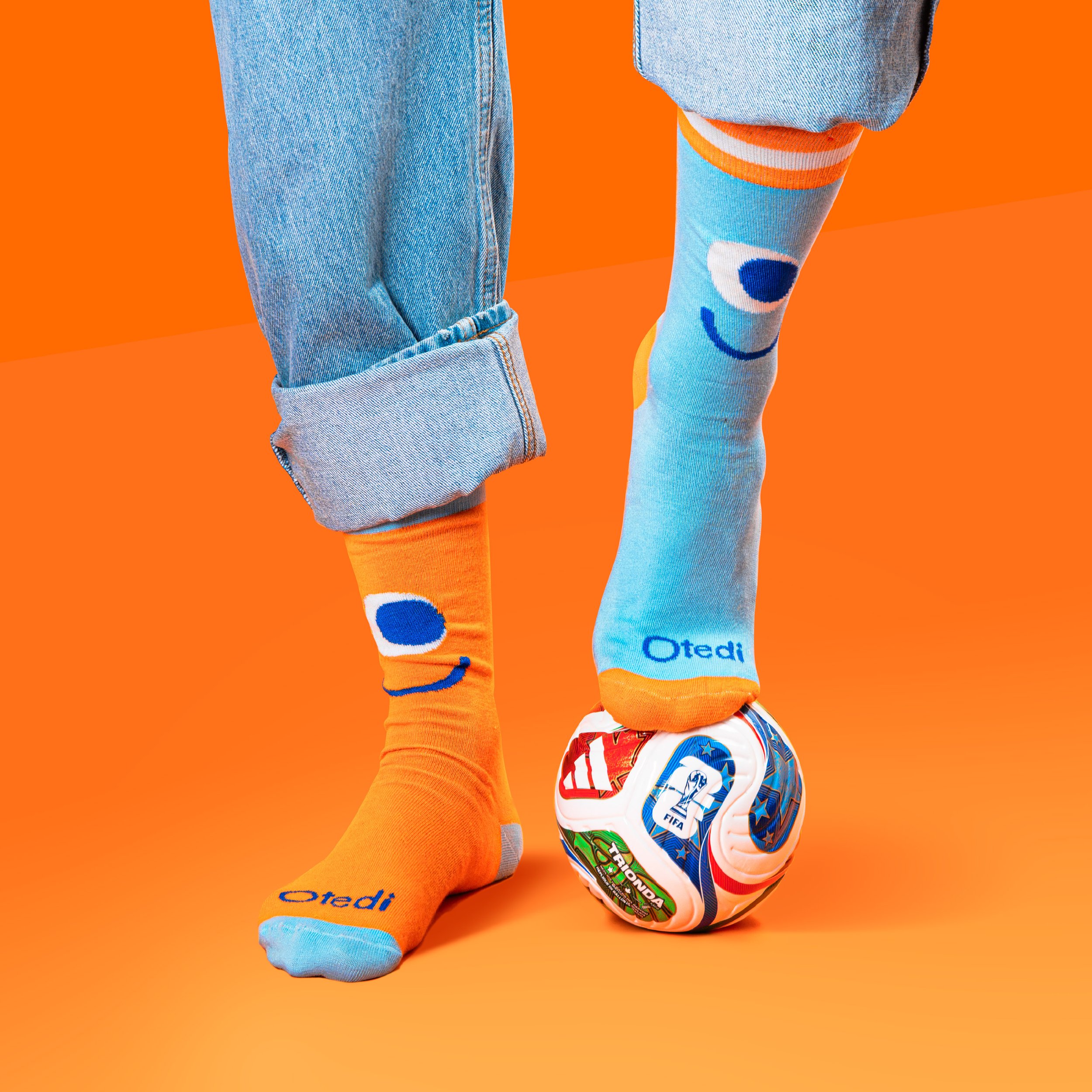 Close-up of two people's legs and feet, one wearing light blue jeans and the other in colorful socks with a face cartoon design, standing on an orange background. Tedi vanguardia en sindrome de down