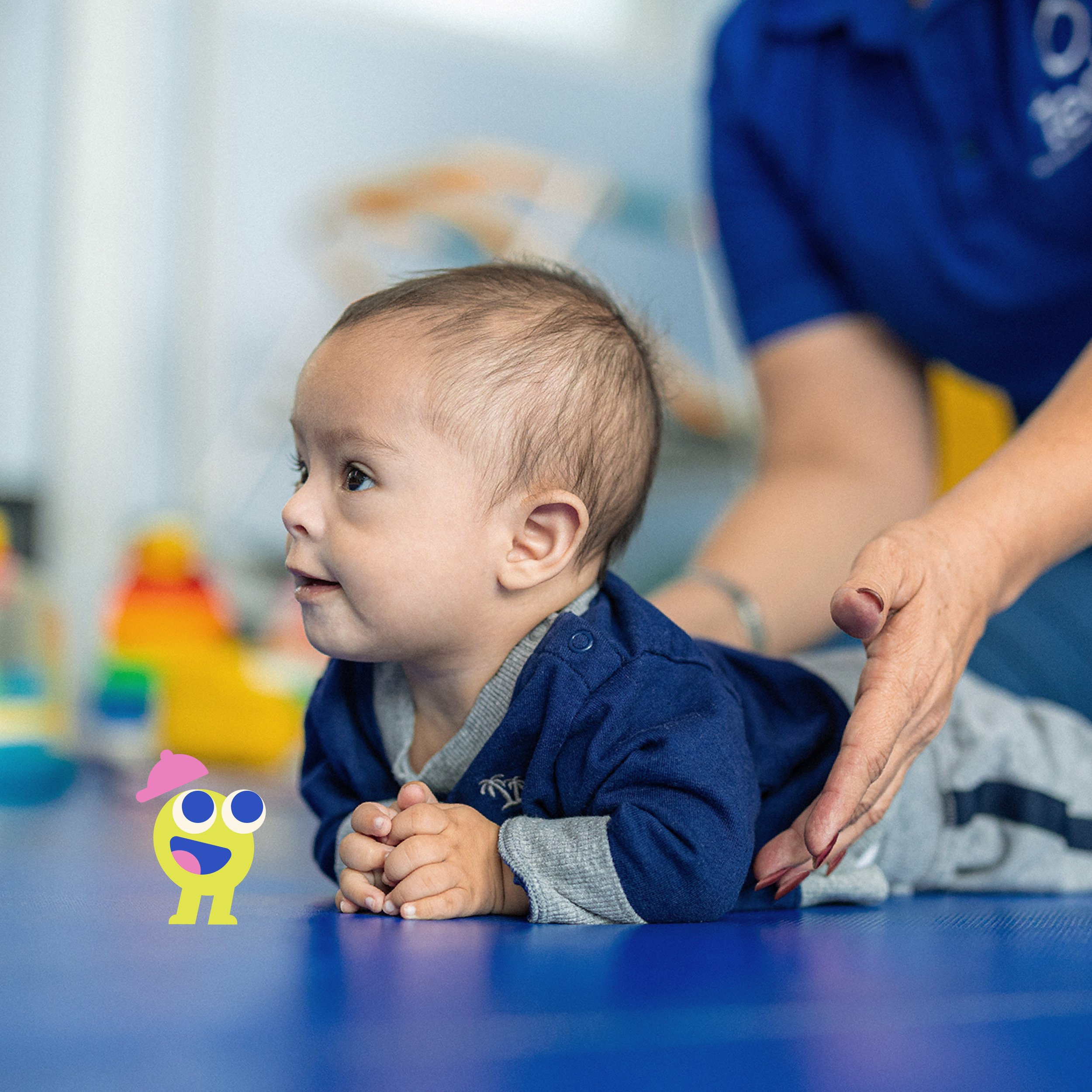A young boy with light skin and short hair is sitting at a blue table, looking to the side with his hands clasped. An adult, wearing red nail polish and a gray sleeve, is gently holding the boy's hands. In the background, there are colorful blurred o