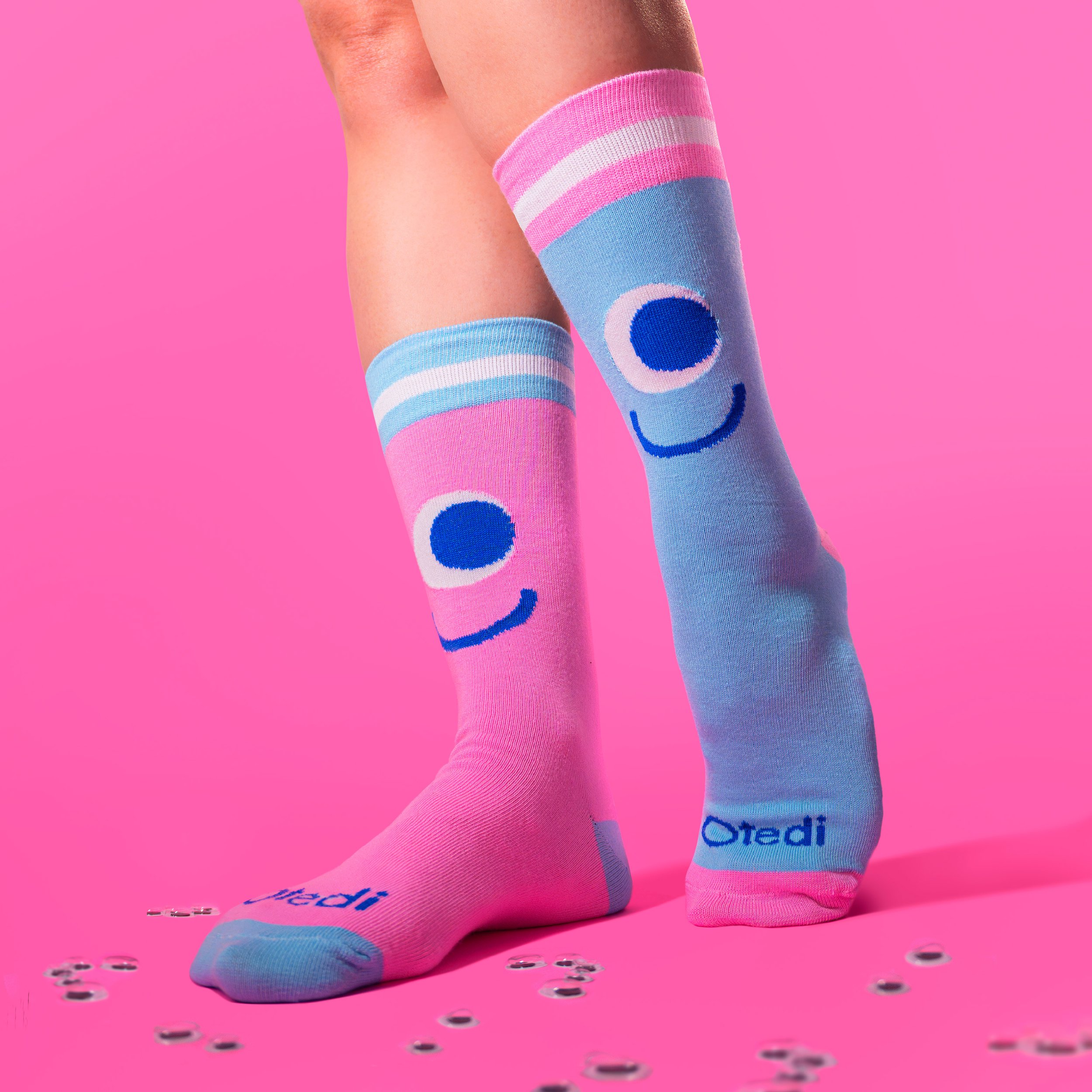 Person wearing colorful knee-high socks with smiley face designs on a pink background, with scattered googly eyes around their feet. Tedi vanguardia en sindrome de down