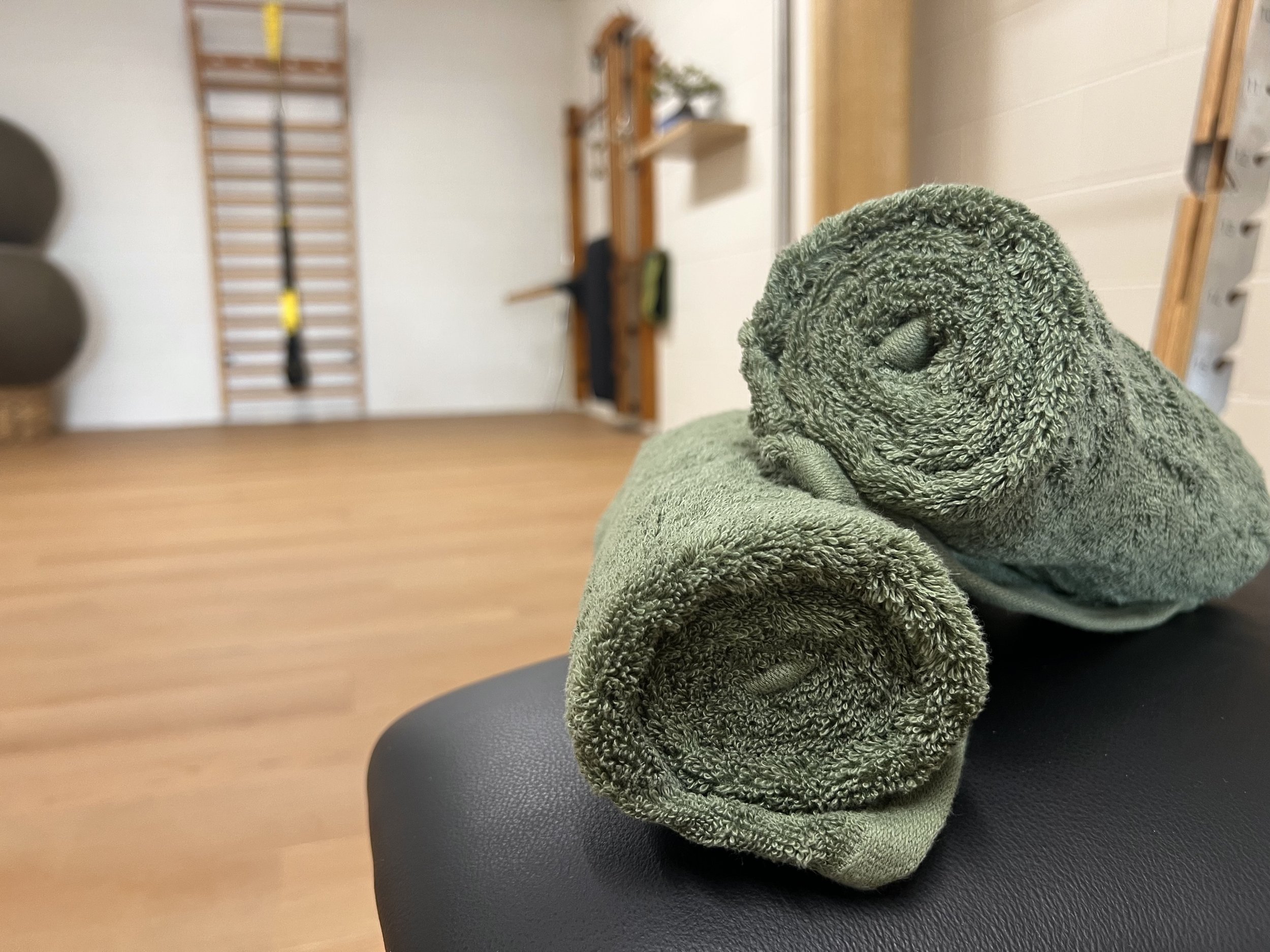 Two green rolled towels on a black surface in a fitness or therapy room with wooden flooring and wall exercise equipment in the background.