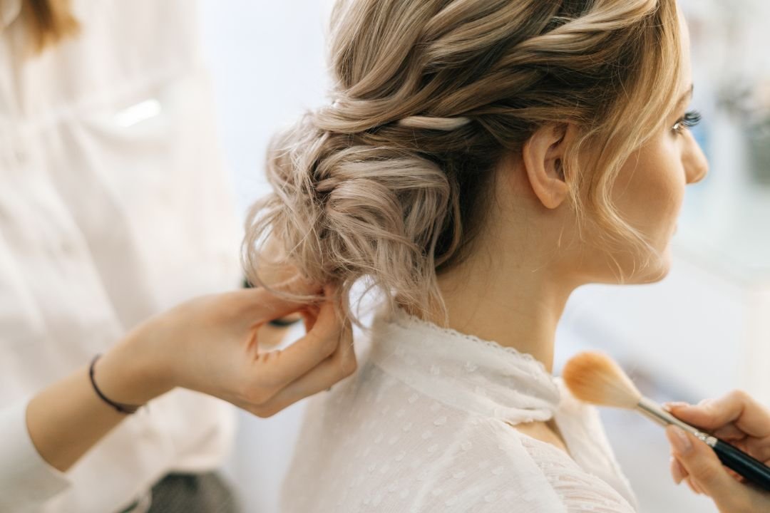 A woman having her hair styled by a hairstylist who is applying makeup with a brush.