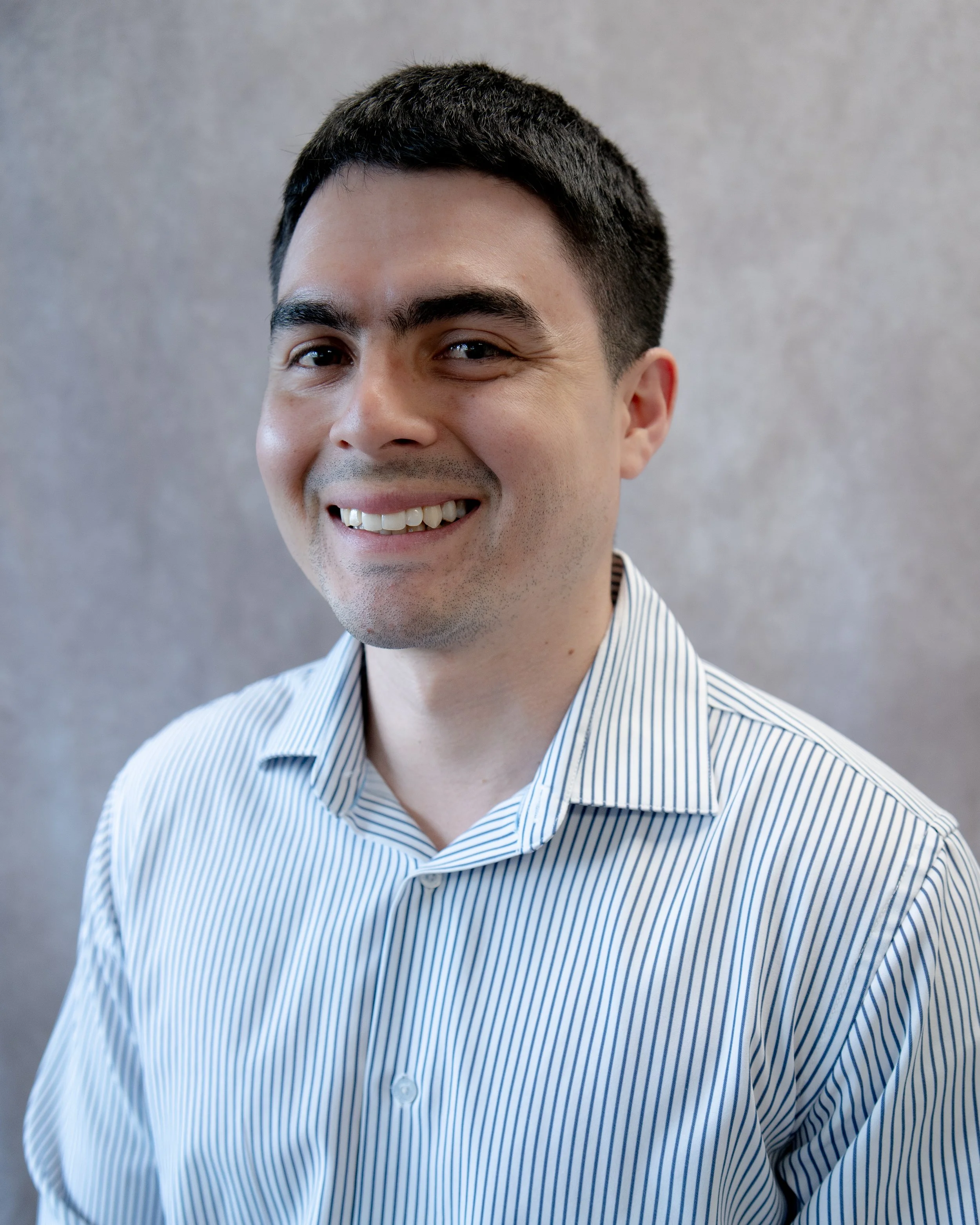 Smiling man with dark hair in a striped dress shirt standing against a neutral background.