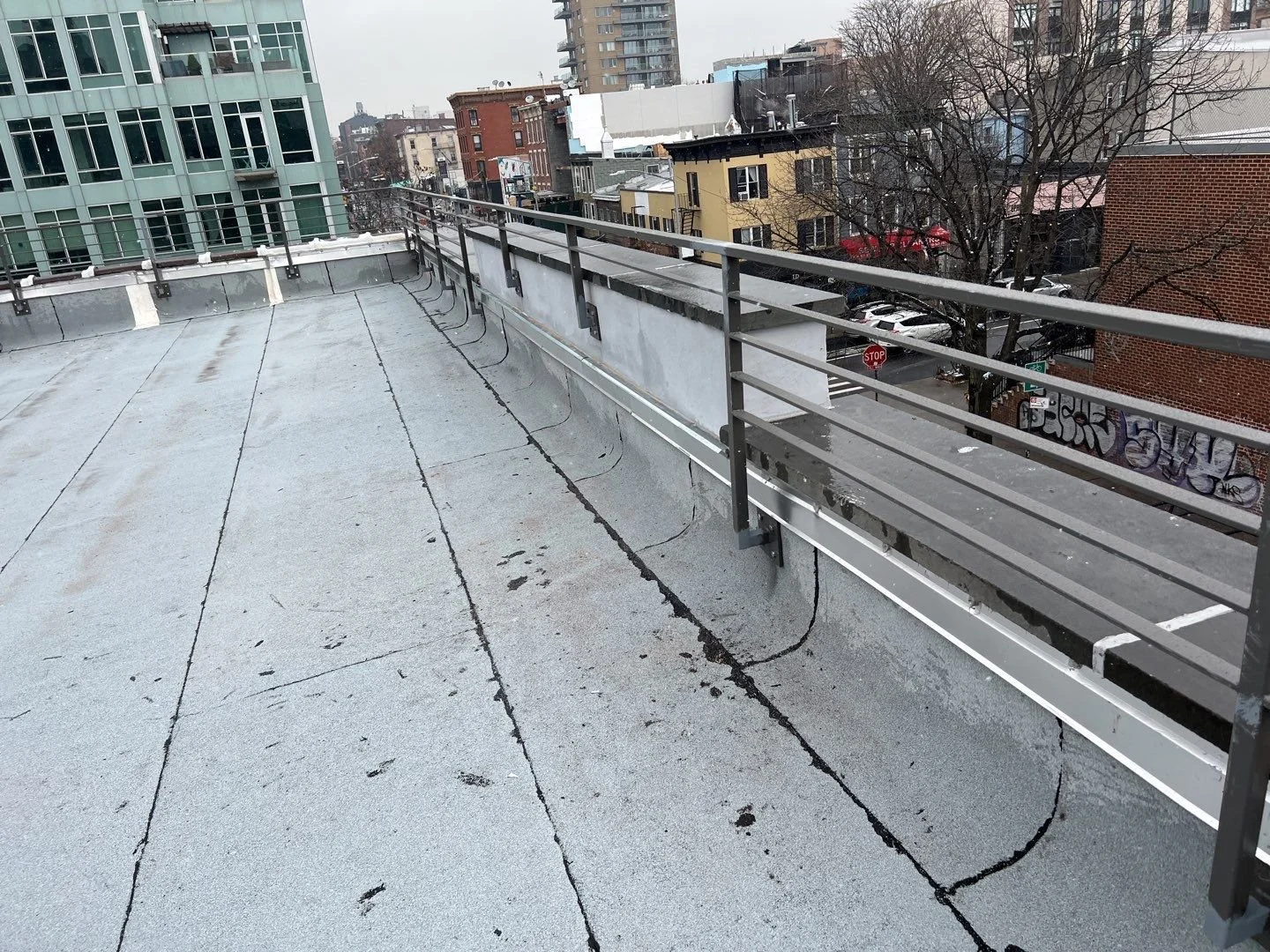 Empty rooftop terrace with black metal railing, city buildings in the background, graffiti on the side of a building, overcast sky.