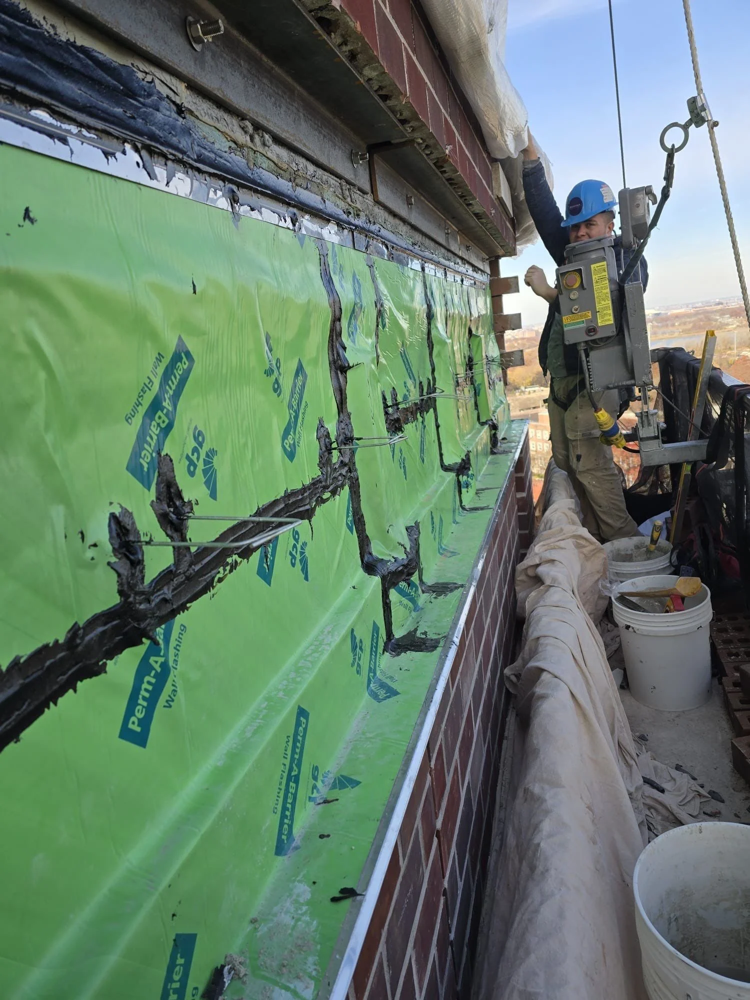Worker applying waterproof sealant or insulation material on a building exterior, wearing a safety helmet and harness, with buckets and tools nearby.