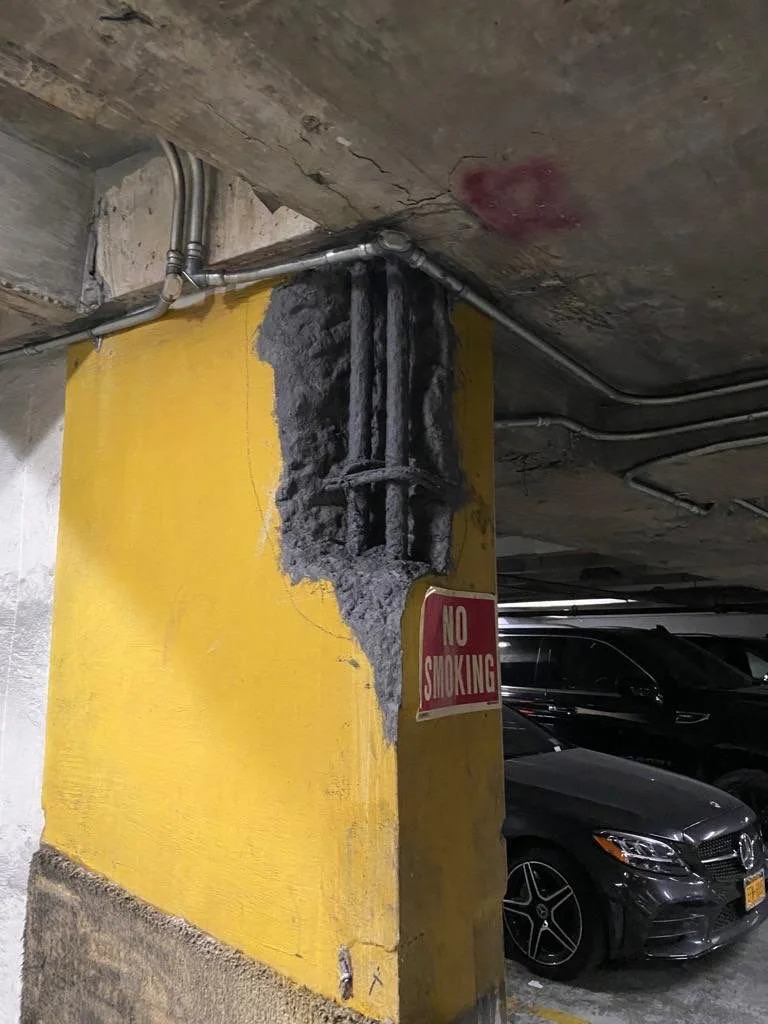Concrete pillar with exposed wiring and a yellow painted side in a parking garage, with a red 'No Smoking' sign attached.