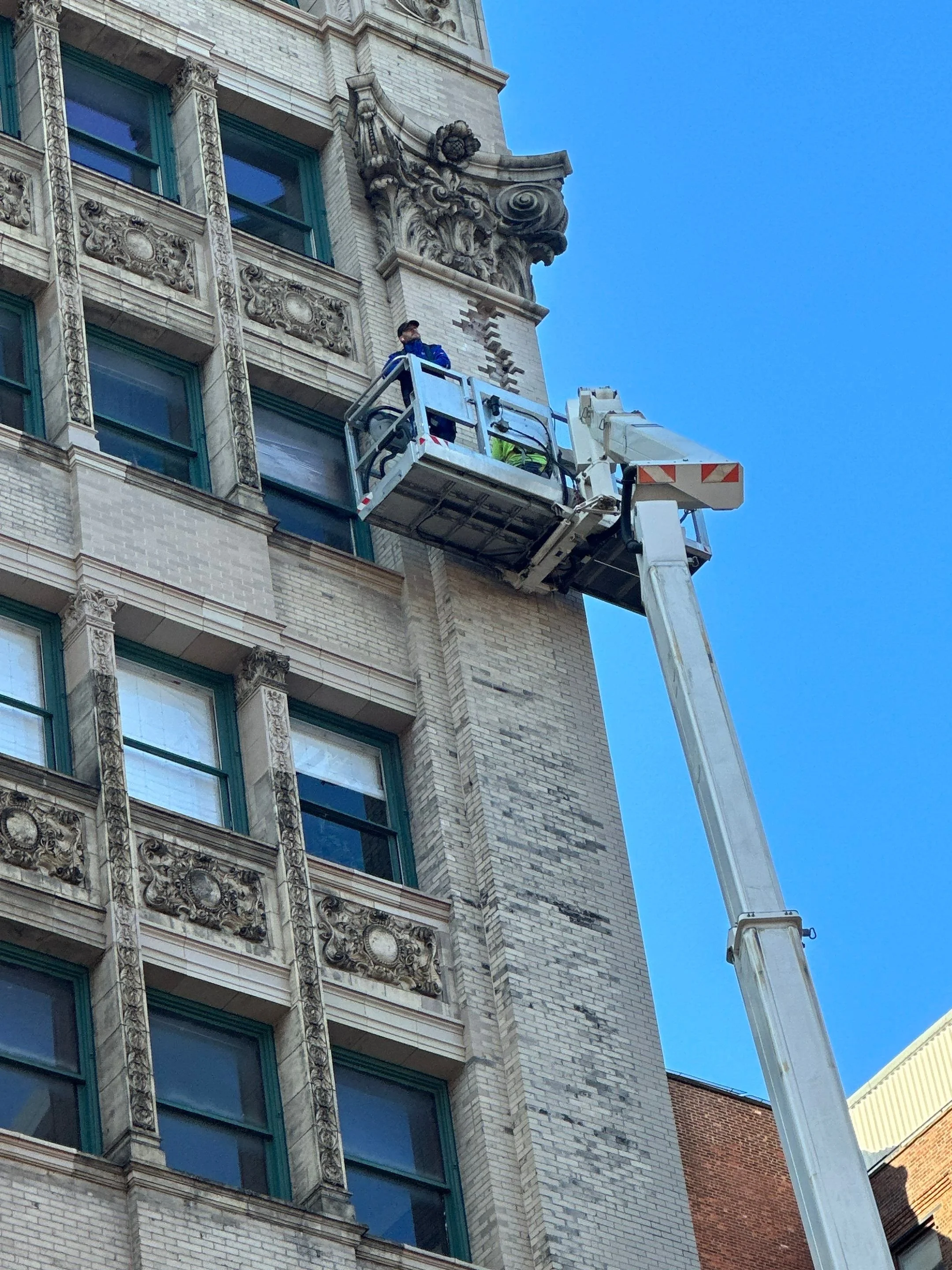 A worker on a cherry picker cleans the ornate architectural details on the corner of a historic building against a clear blue sky.