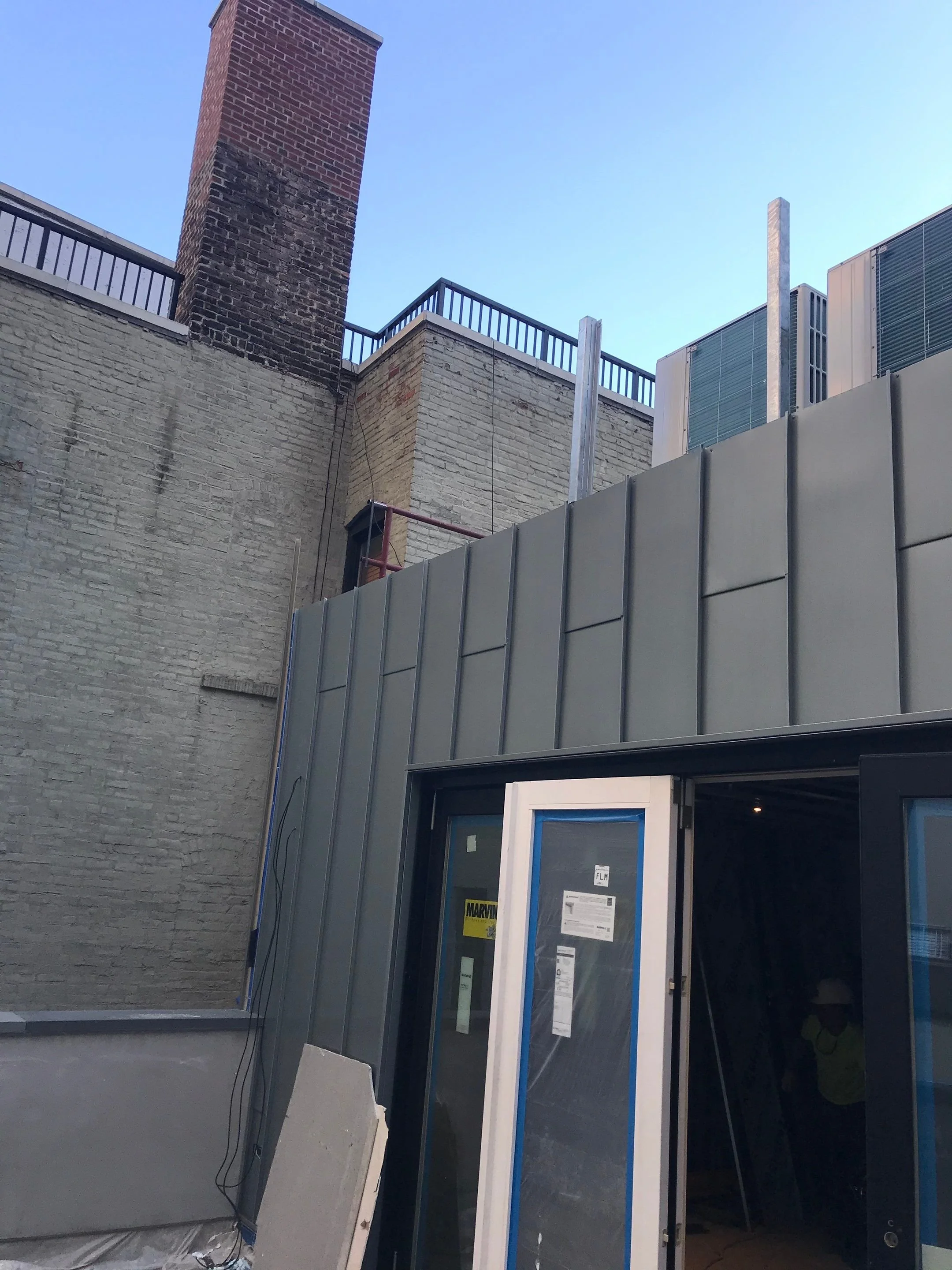 Construction site with a building under renovation, featuring a metal exterior, large door, and high brick walls with a chimney and railing on top.