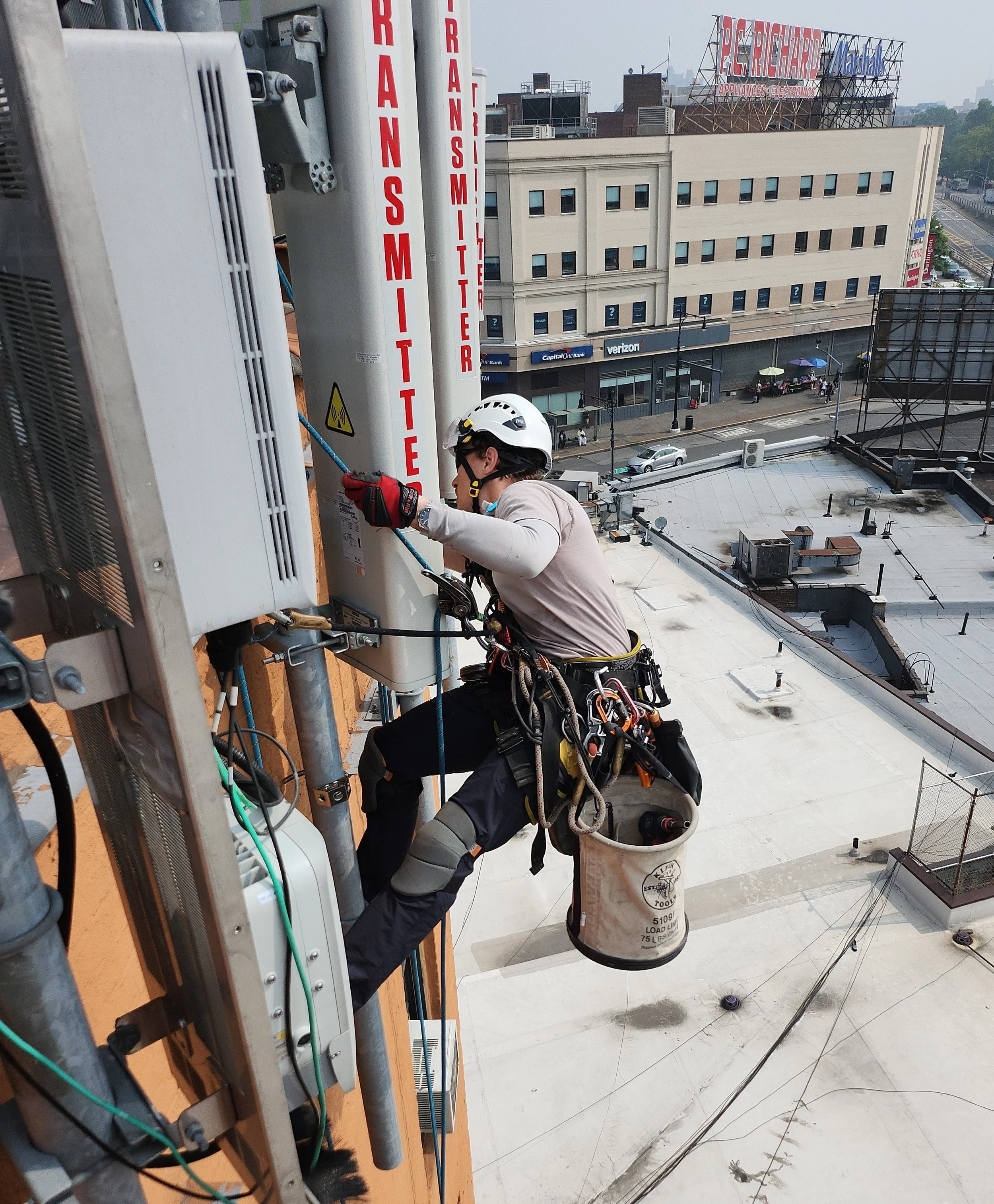 A construction worker wearing a harness and helmet is working on telecommunications equipment on the side of a building, with city streets and buildings visible in the background.