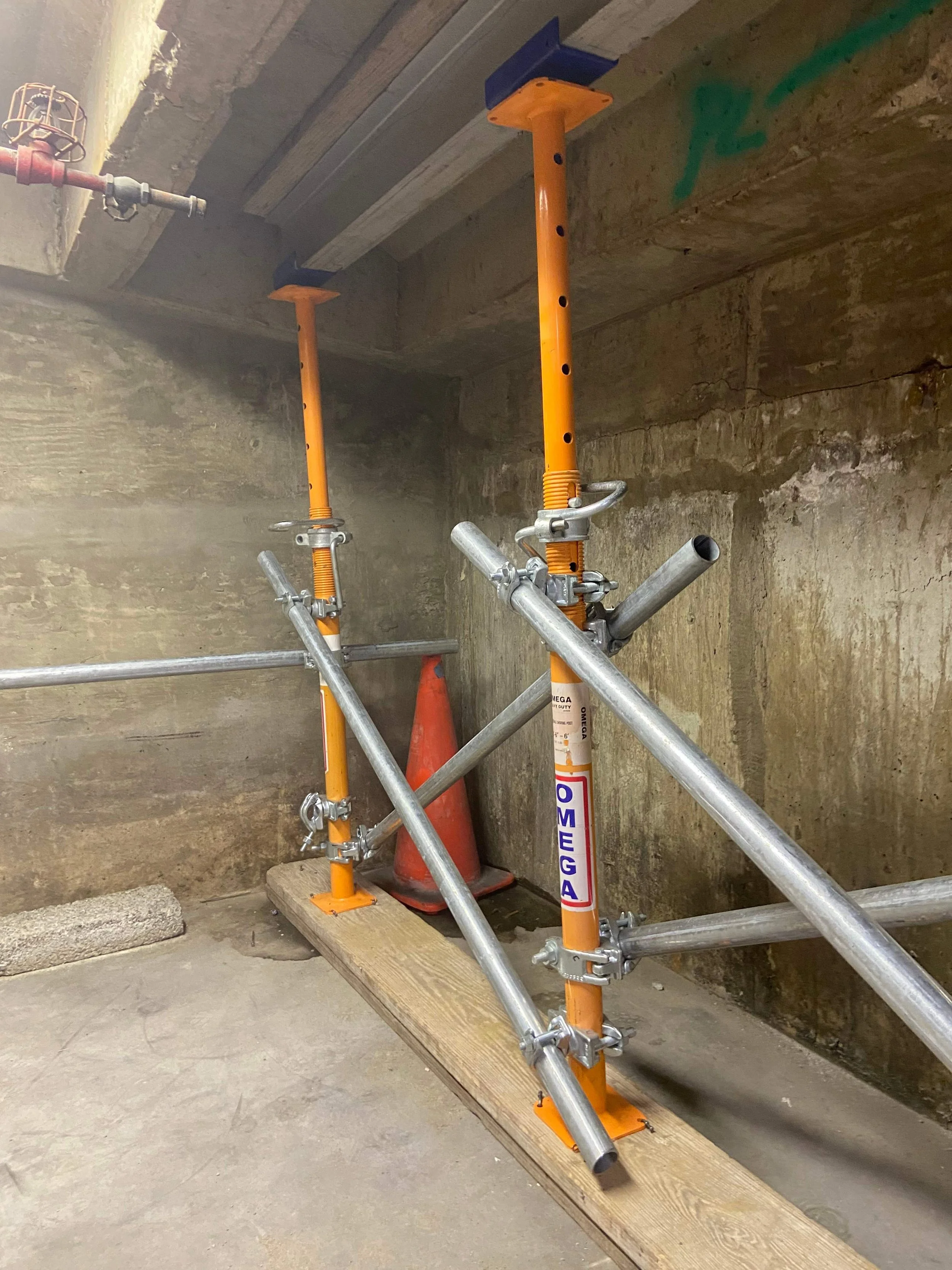 Two orange and silver adjustable scaffolding poles with the label "OMEGA" in a construction area, positioned on a wooden plank inside a concrete room.