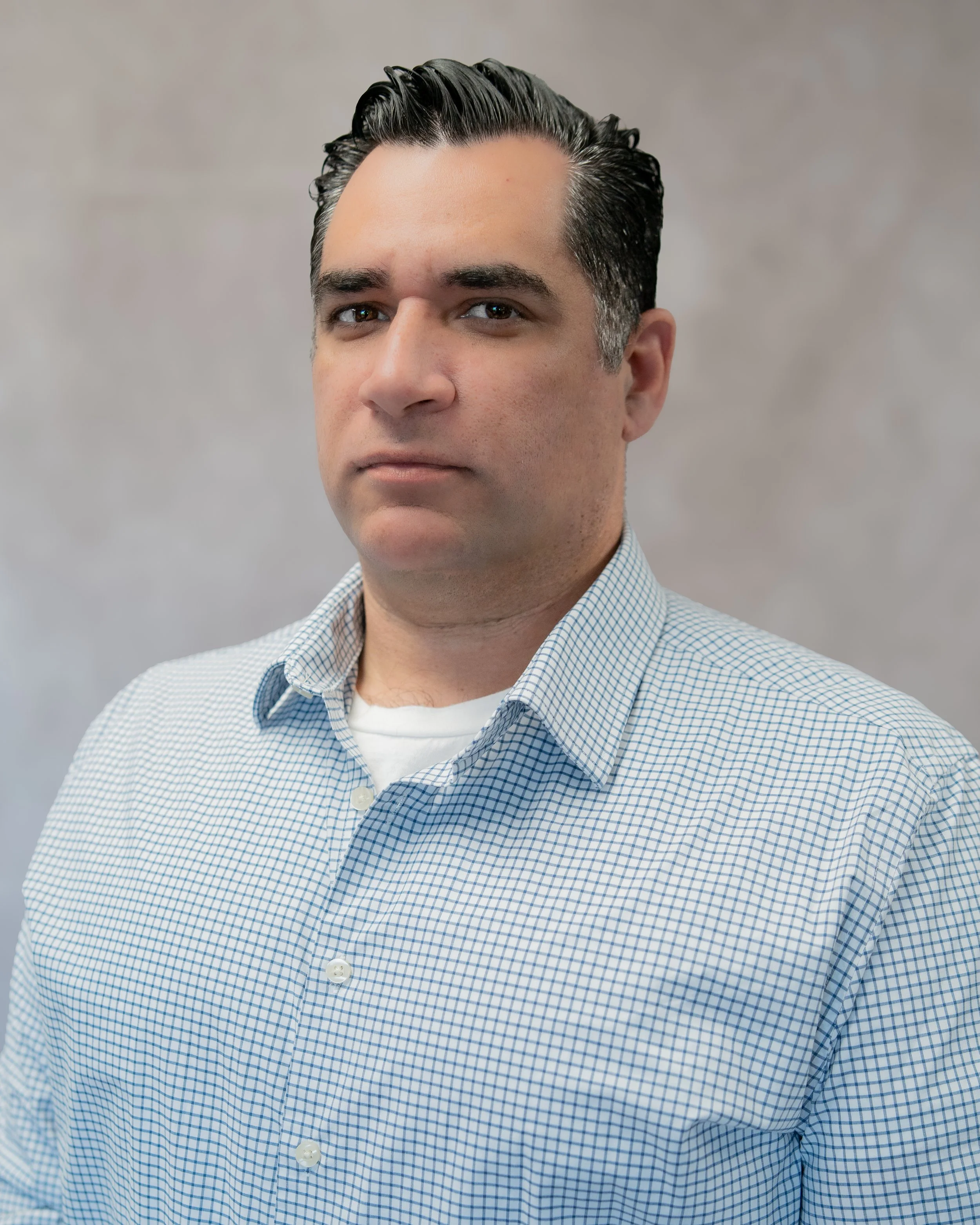 Portrait of a man with dark, slicked-back hair wearing a light blue checkered shirt and a white undershirt against a neutral background.
