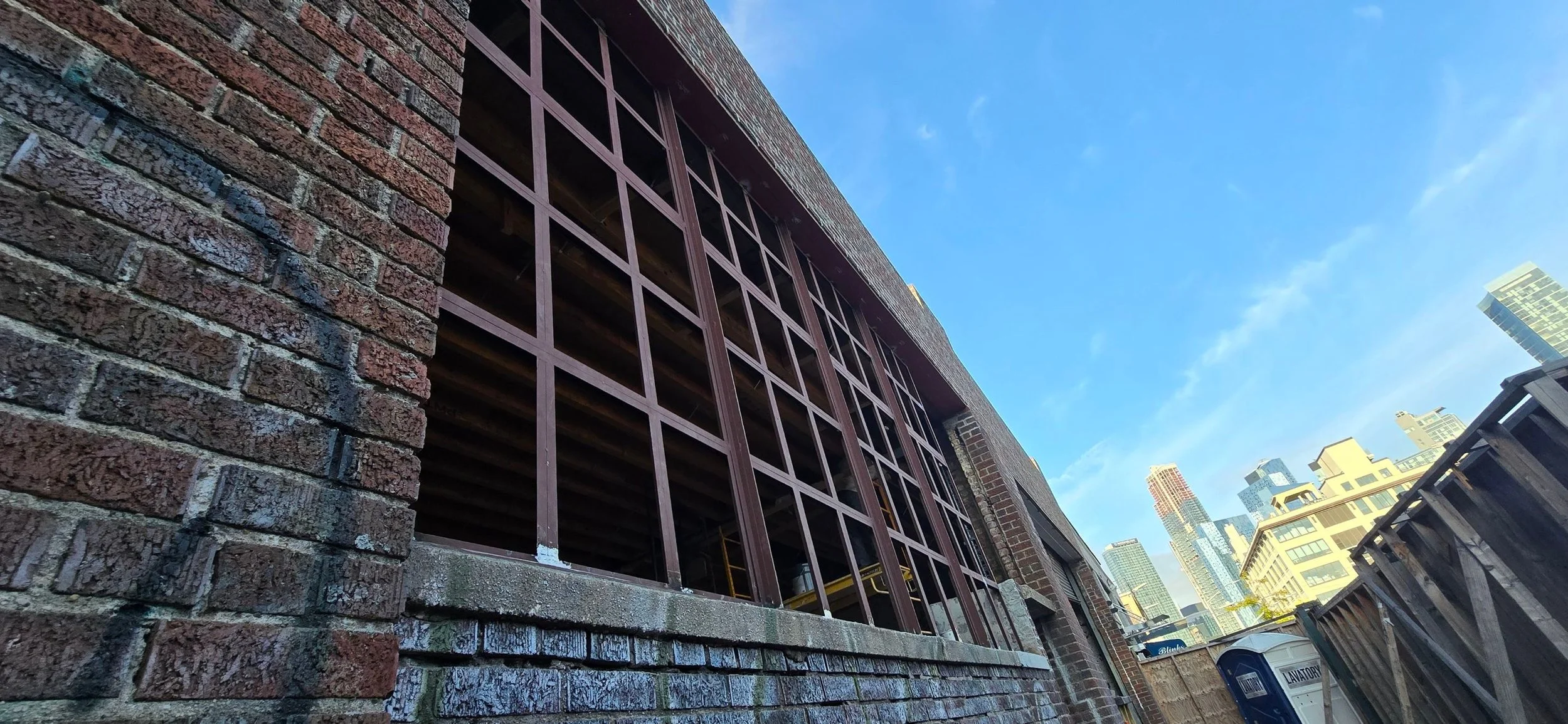 View of a brick building under construction with a large window frame and a city skyline in the background on a clear day.