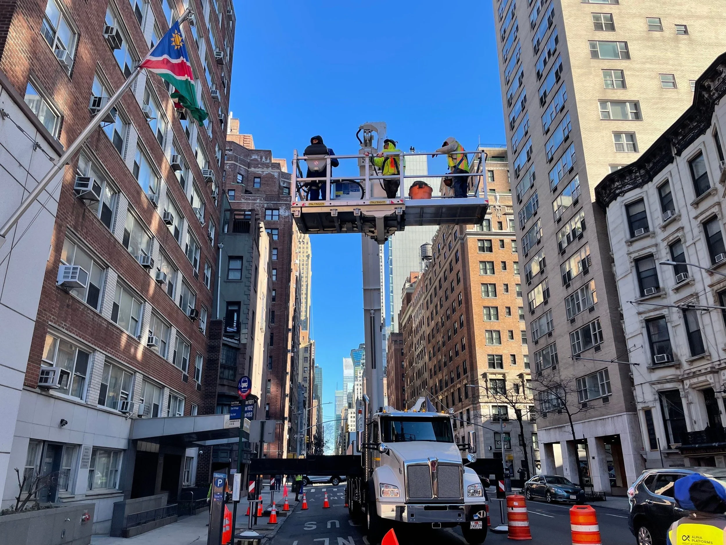 A city street scene with workers on an elevated platform repairing or installing something on the building at a height, with tall buildings on either side and orange traffic cones on the street.