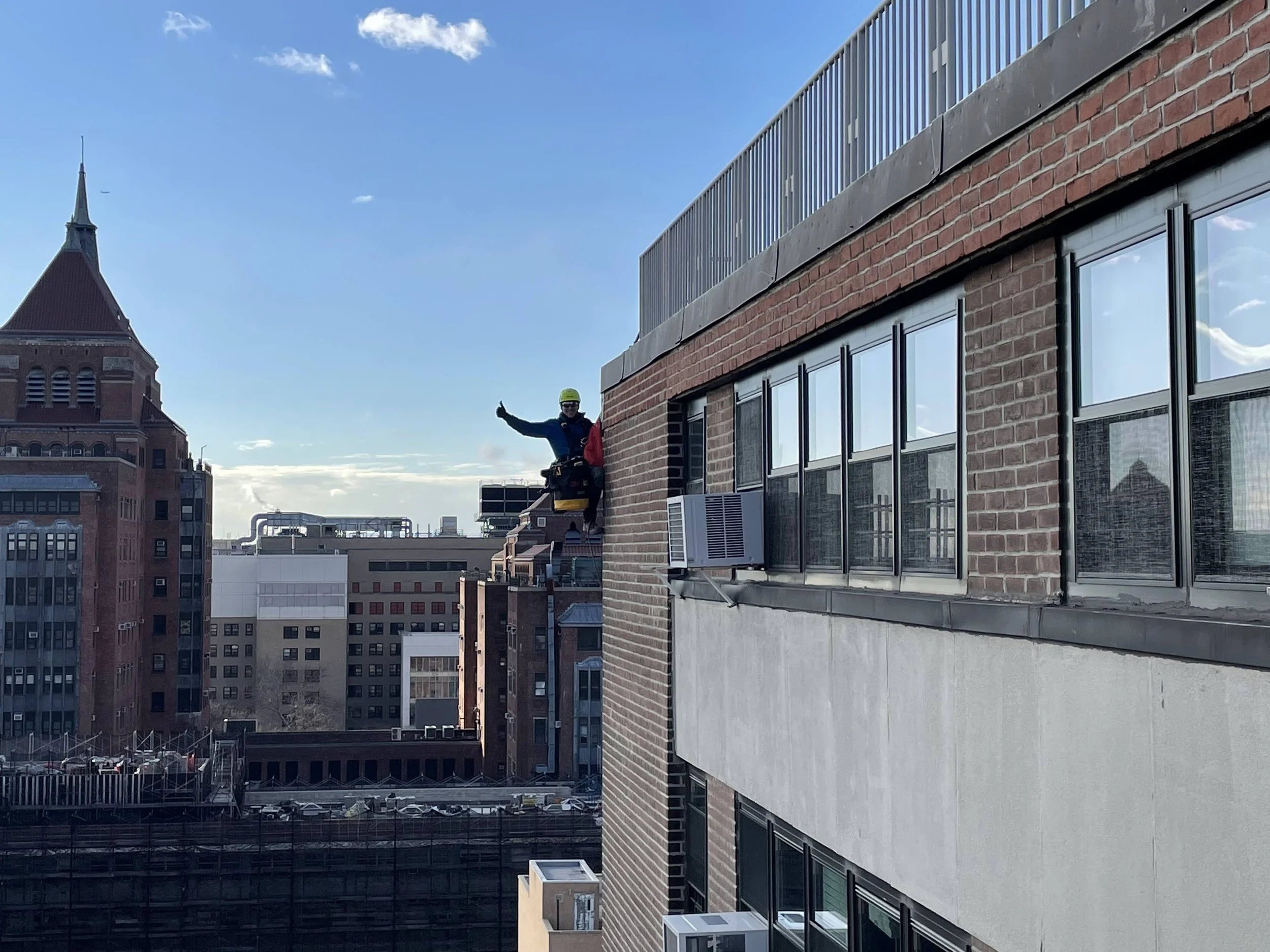 A person wearing a yellow helmet, sunglasses, and a safety harness is sitting on the edge of the upper floor of a brick building, giving a thumbs-up and waving, with a cityscape of tall buildings and a bright blue sky in the background.