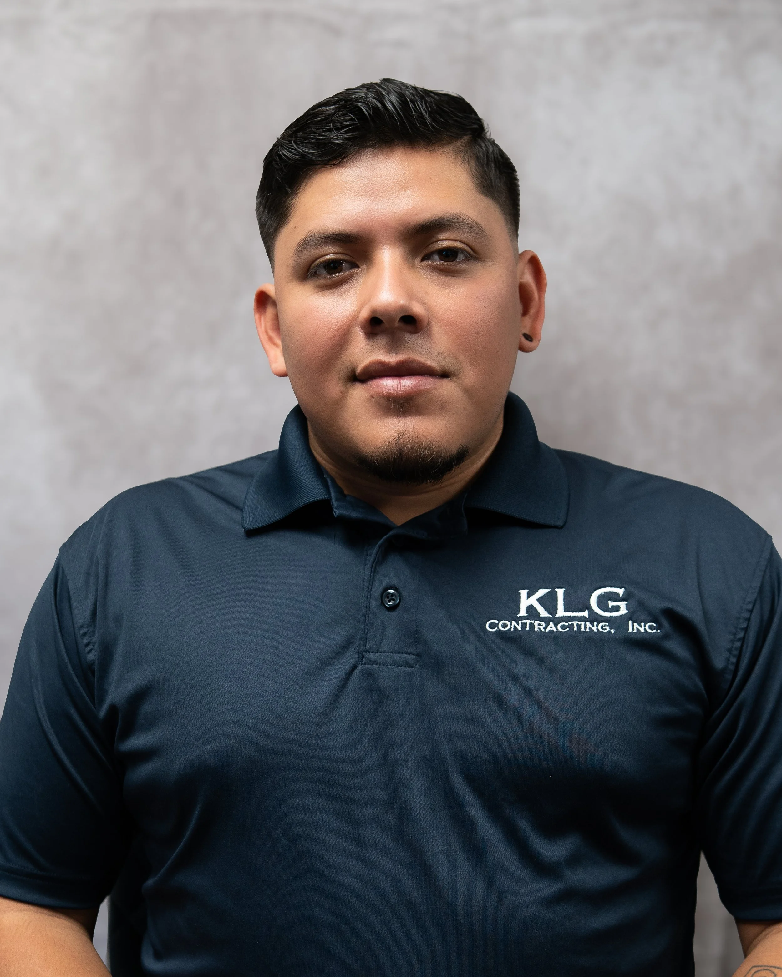A young man with dark hair and a goatee wearing a navy blue polo shirt with the logo 'KLG Contracting, Inc.' stands against a gray background.