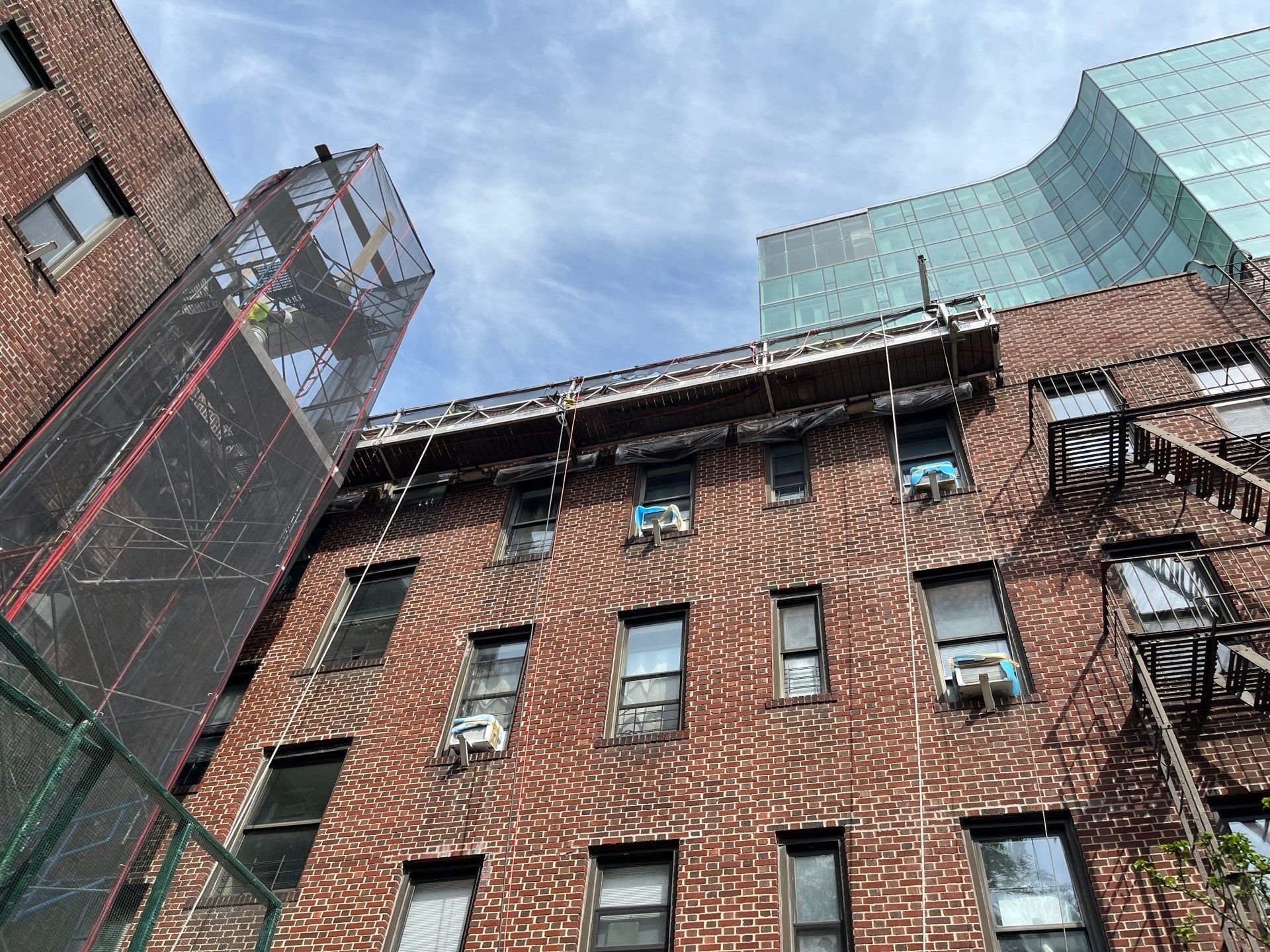 A multi-story brick building with several windows, some with air conditioning units. Scaffolding and construction netting are visible on parts of the building, with a modern glass structure attached to the top. The sky is blue with some clouds.
