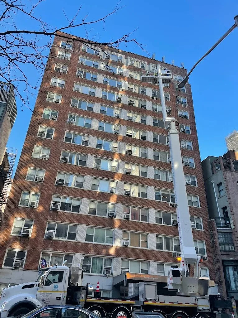 A tall brick apartment building with multiple windows, some with air conditioning units. A cherry tree branch extends into the frame at the top. A utility bucket truck is parked in front, with a worker inside the bucket near the building, likely performing maintenance or repairs. The sky is clear and blue.