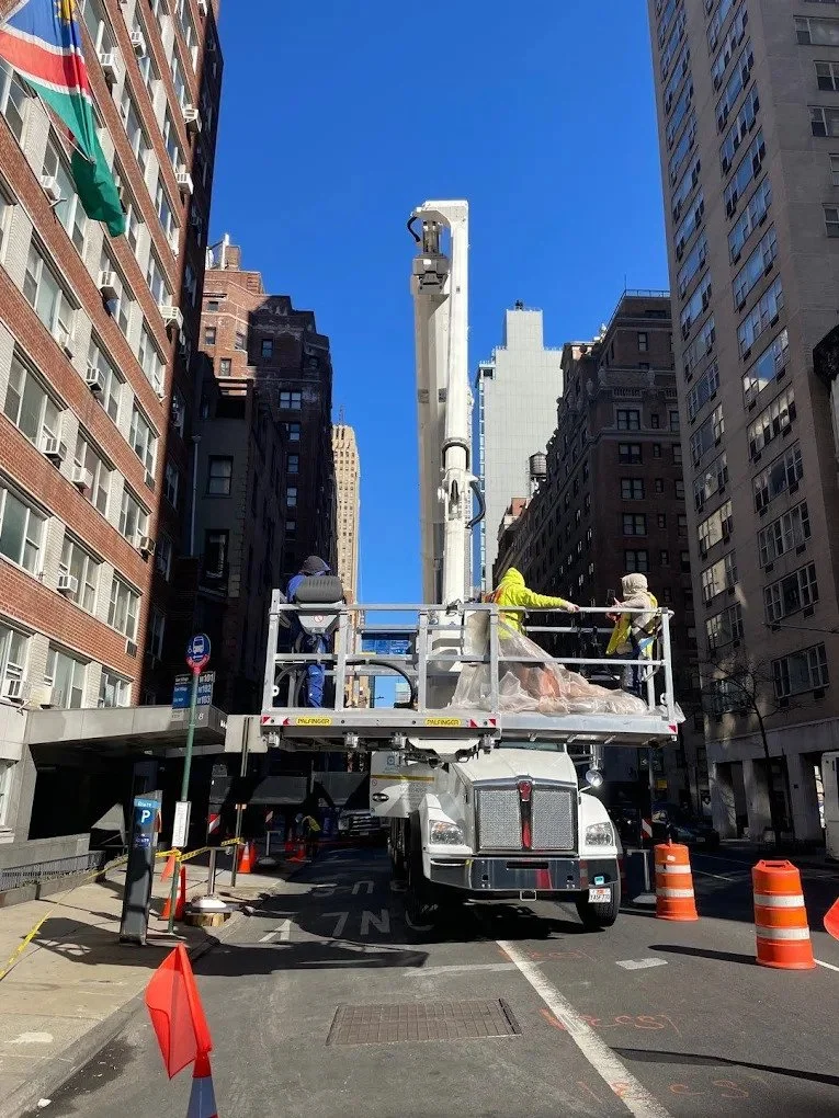 Construction workers on a lift working on a tall street light or utility pole in an urban city street surrounded by tall buildings, with traffic cones, and a blue sky.