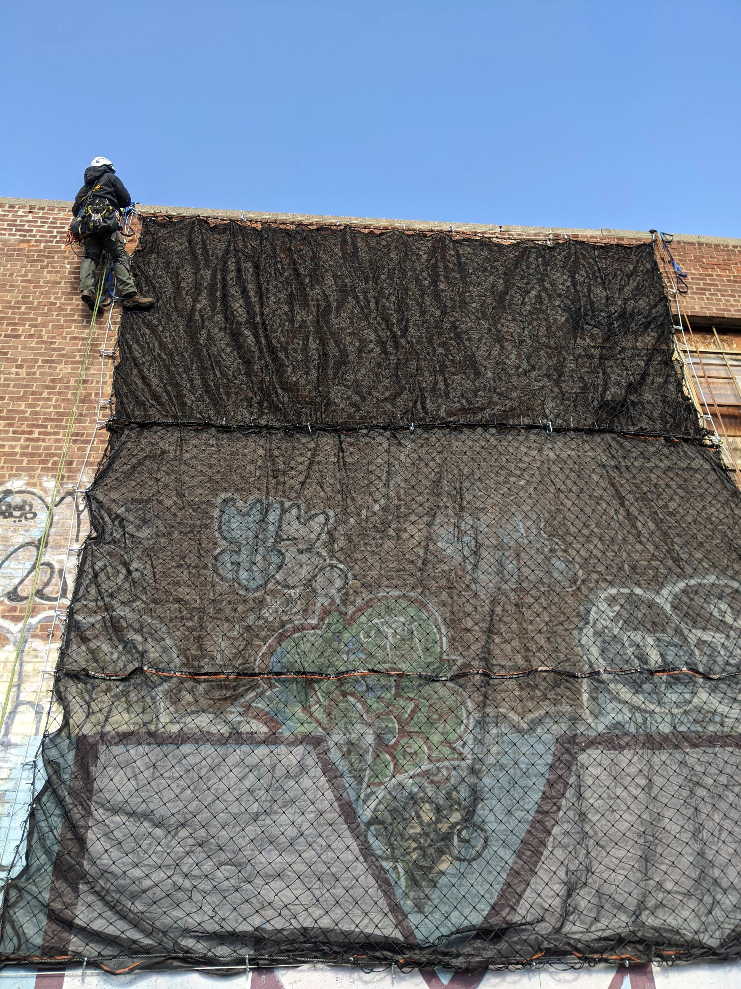 A worker in safety gear climbing a ladder attached to a building with graffiti-covered wall behind a netting.