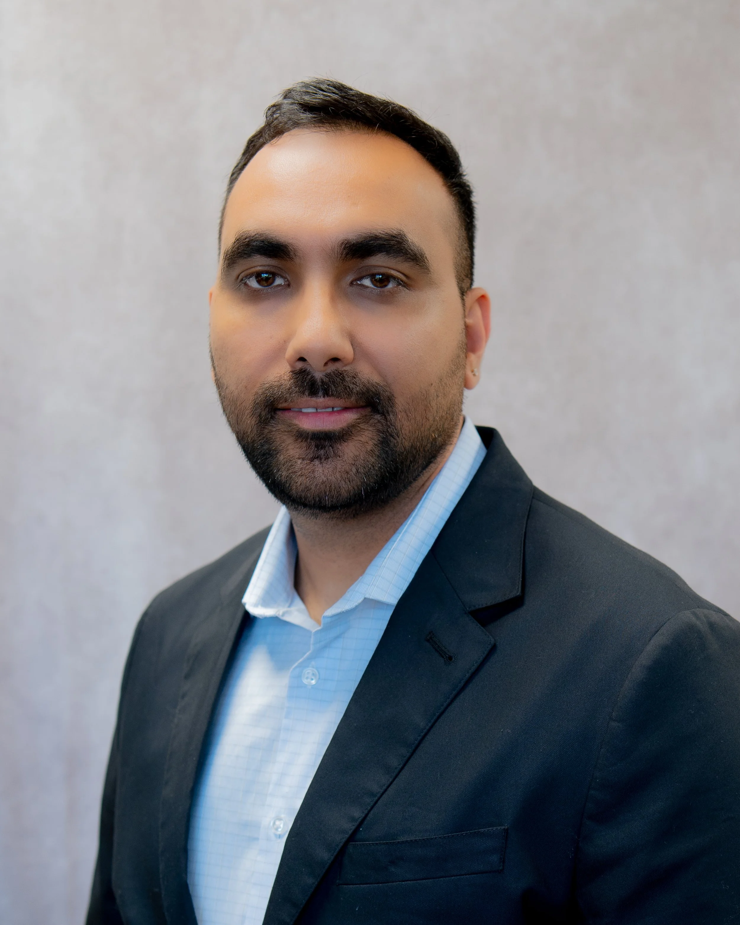 Professional headshot of a man with dark hair, beard, wearing a black blazer and a light blue shirt, standing against a neutral background.