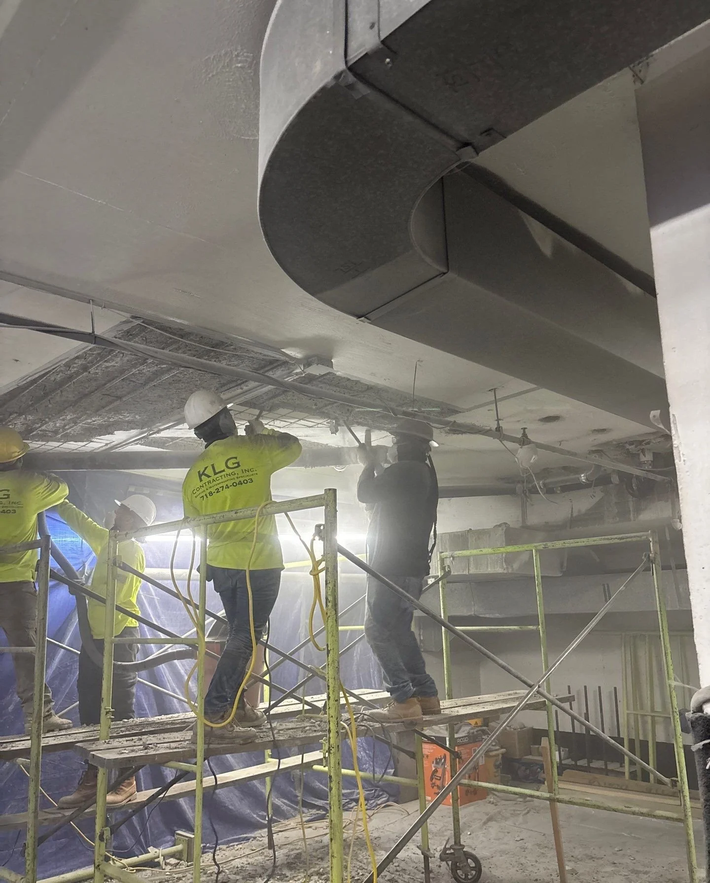 Construction workers on scaffolding working on ceiling pipes in an indoor construction site.
