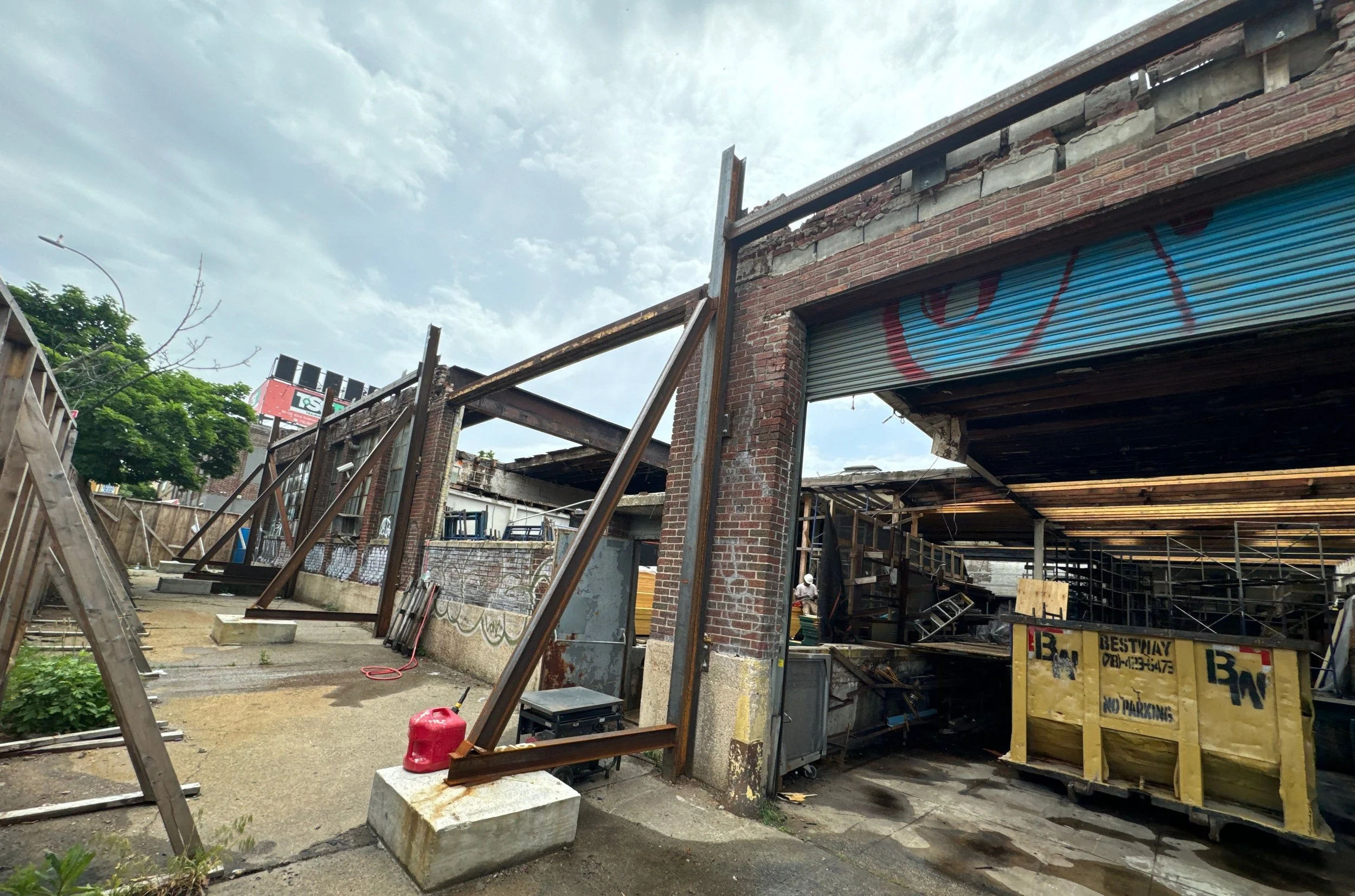 An urban construction site with a partially dismantled brick building, scaffolding, and a yellow barricade. There is a red gas can, a ladder, and a blue sky with some clouds.