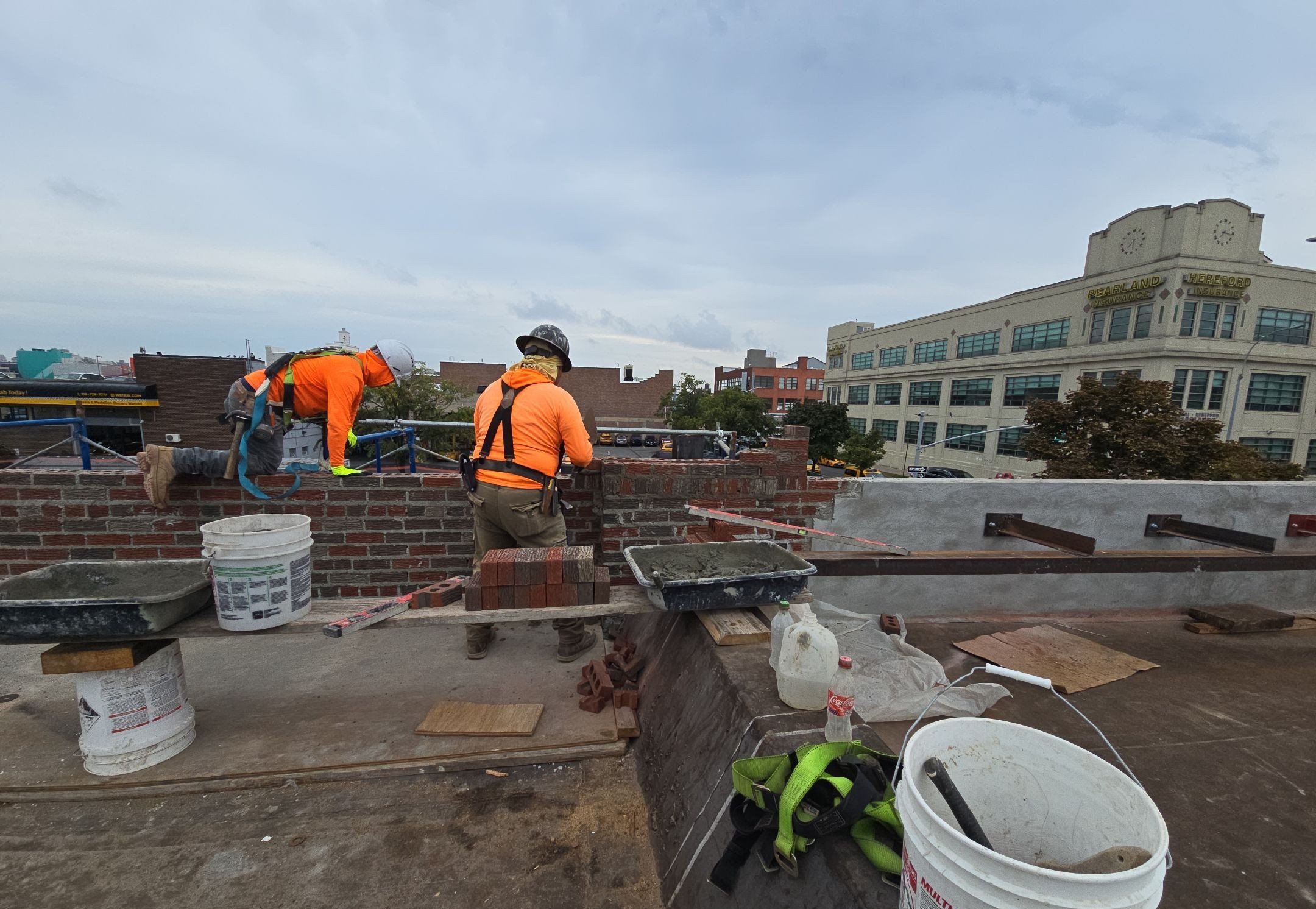 Construction workers wearing orange safety jackets and helmets working on a building rooftop, with tools, buckets, and bricks around them, and city buildings in the background.