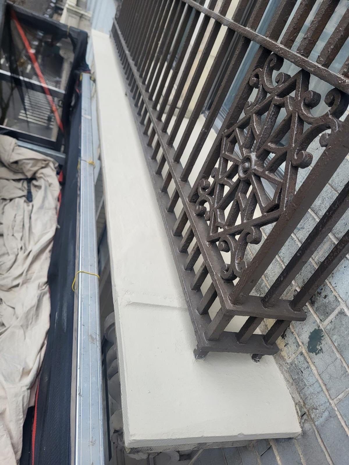 Close-up of a small black metal decorative railing on a beige concrete base, next to a brick wall and a construction area with tools and materials.