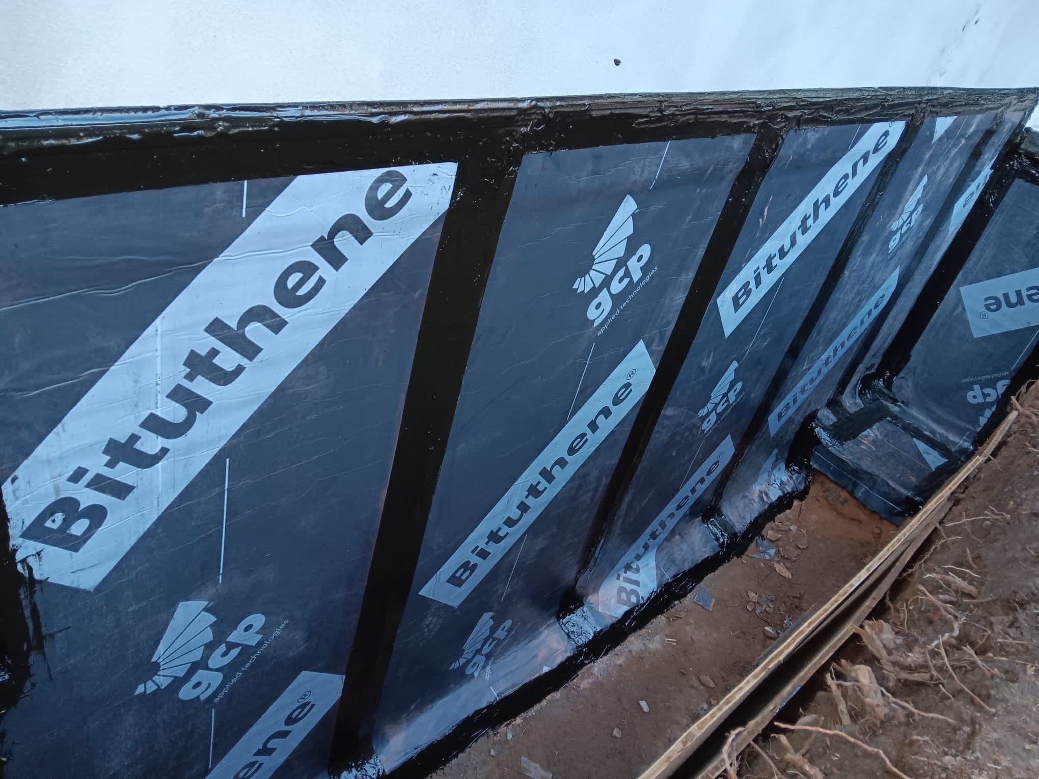 Close-up of a building's exterior wall under construction, covered with black siding or insulation material labeled 'Bituthene' from GCP and 'Biturtherm'.