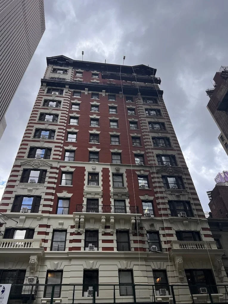 Tall historic building with ornate details, red brick and cream-colored accents, and windows with air conditioning units, under a cloudy sky.