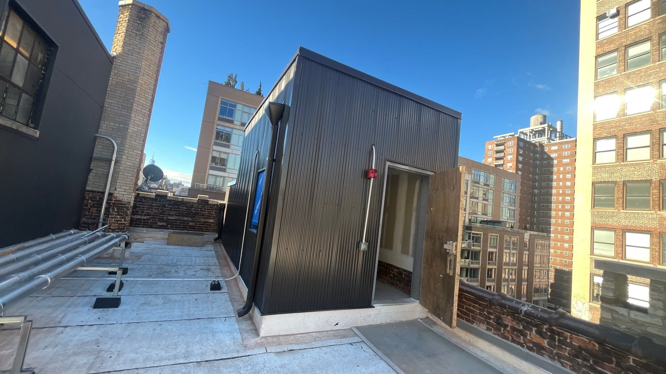 Rooftop with small black metal structure, electrical conduit, and cityscape background with high-rise buildings under a blue sky.