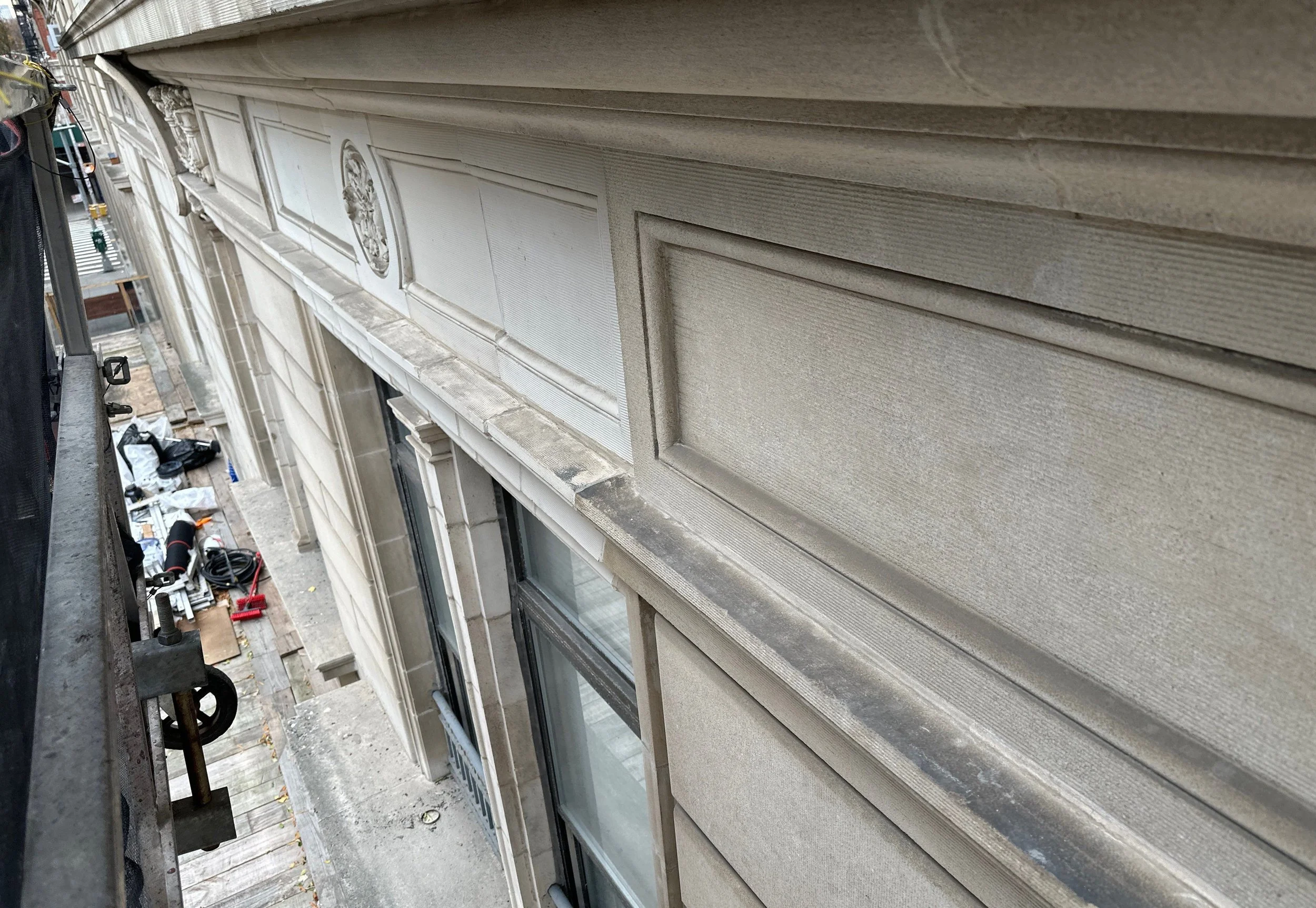 View of the side of a building showing intricate stonework and large windows, with a small balcony and various tools and materials on the ground below, seen from an upward angle.