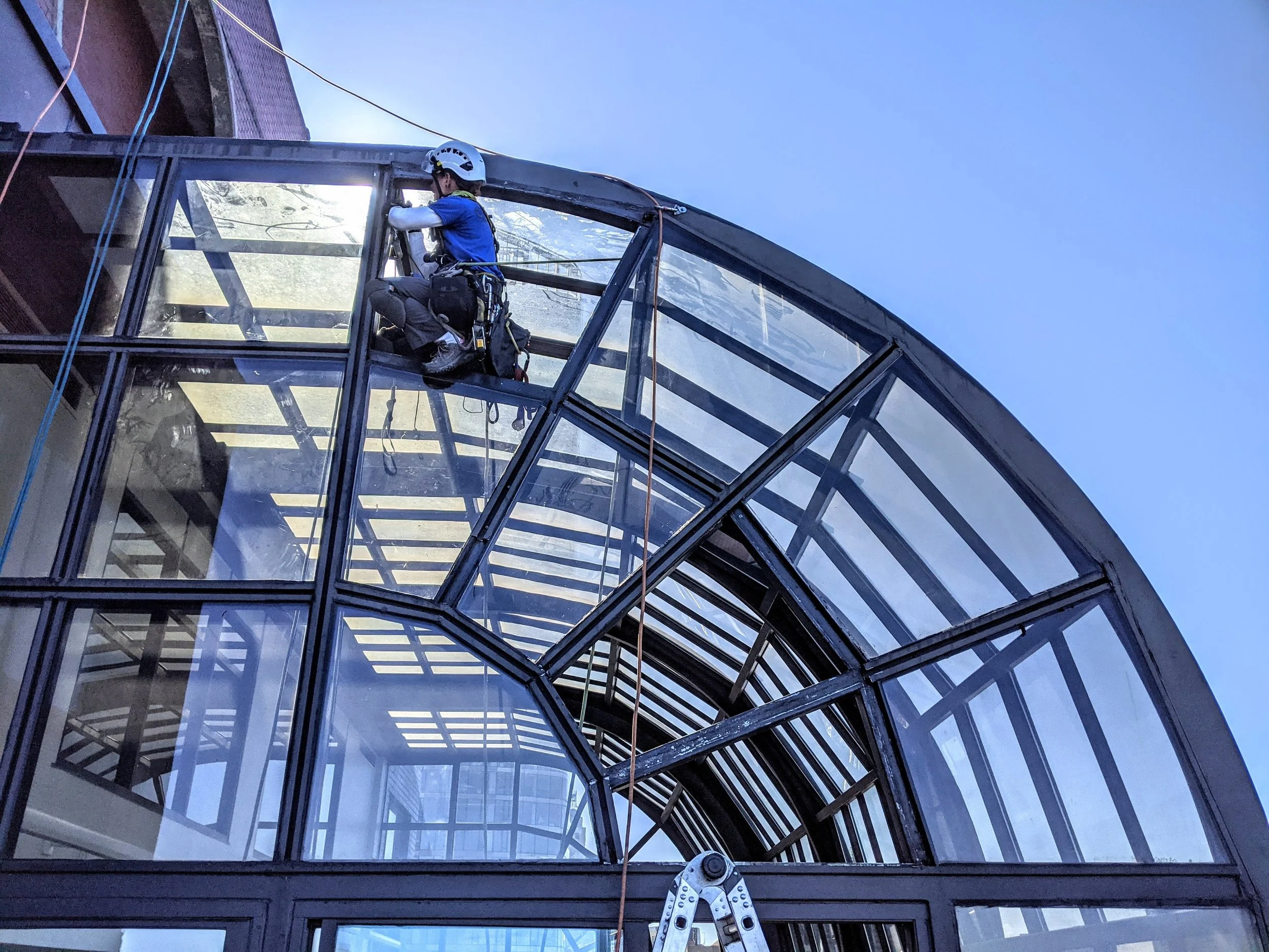 A worker wearing safety gear and a helmet is cleaning or inspecting the glass roof of a building using a harness, ropes, and a ladder.
