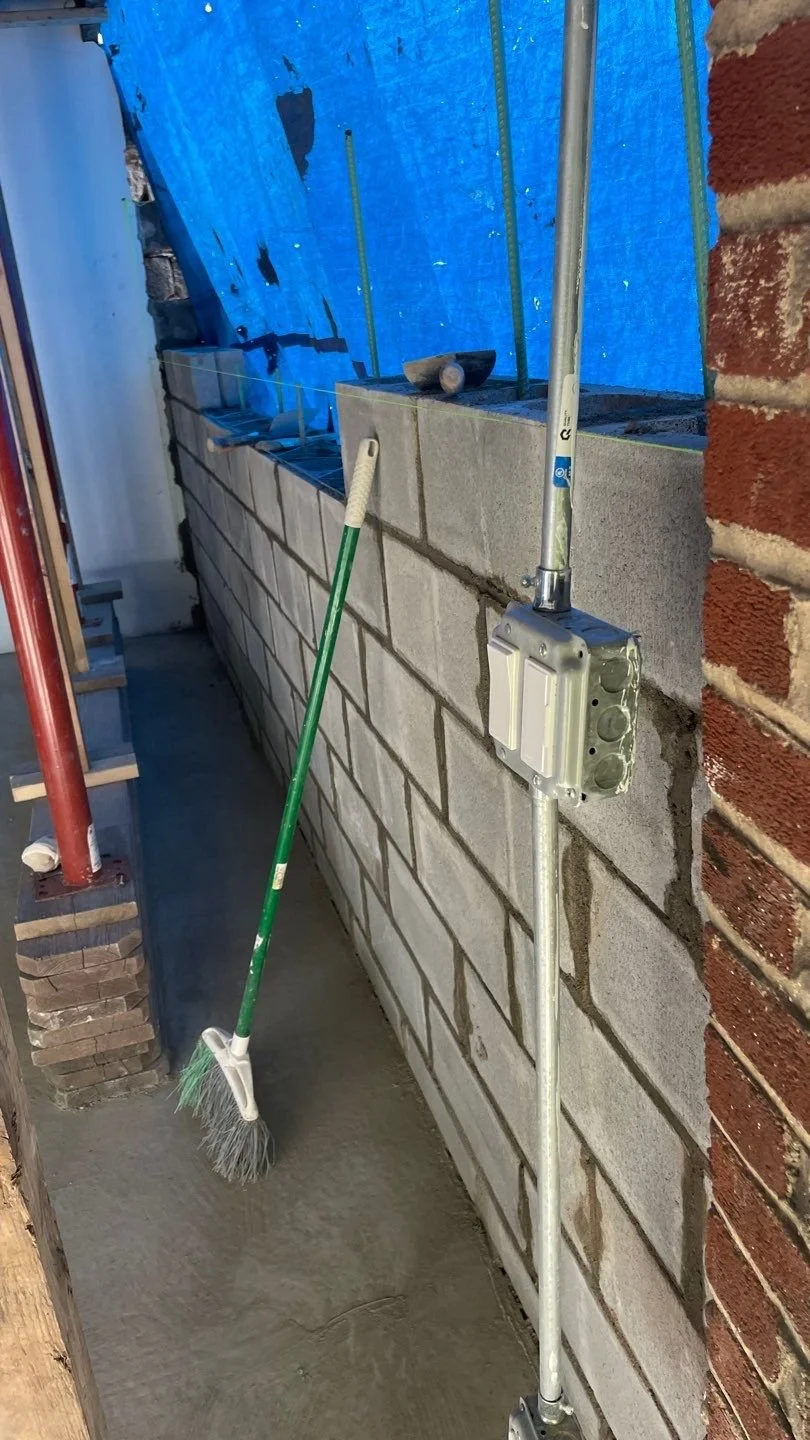 Construction site with brick wall and cinder block wall, construction tools including a broom leaning against the wall, and a blue tarp covering part of the area.