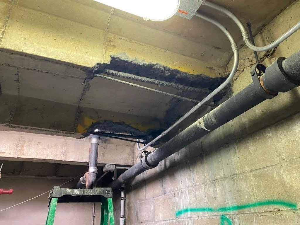 Ceiling with exposed pipes and electrical wiring, some concrete patch work and a ladder underneath