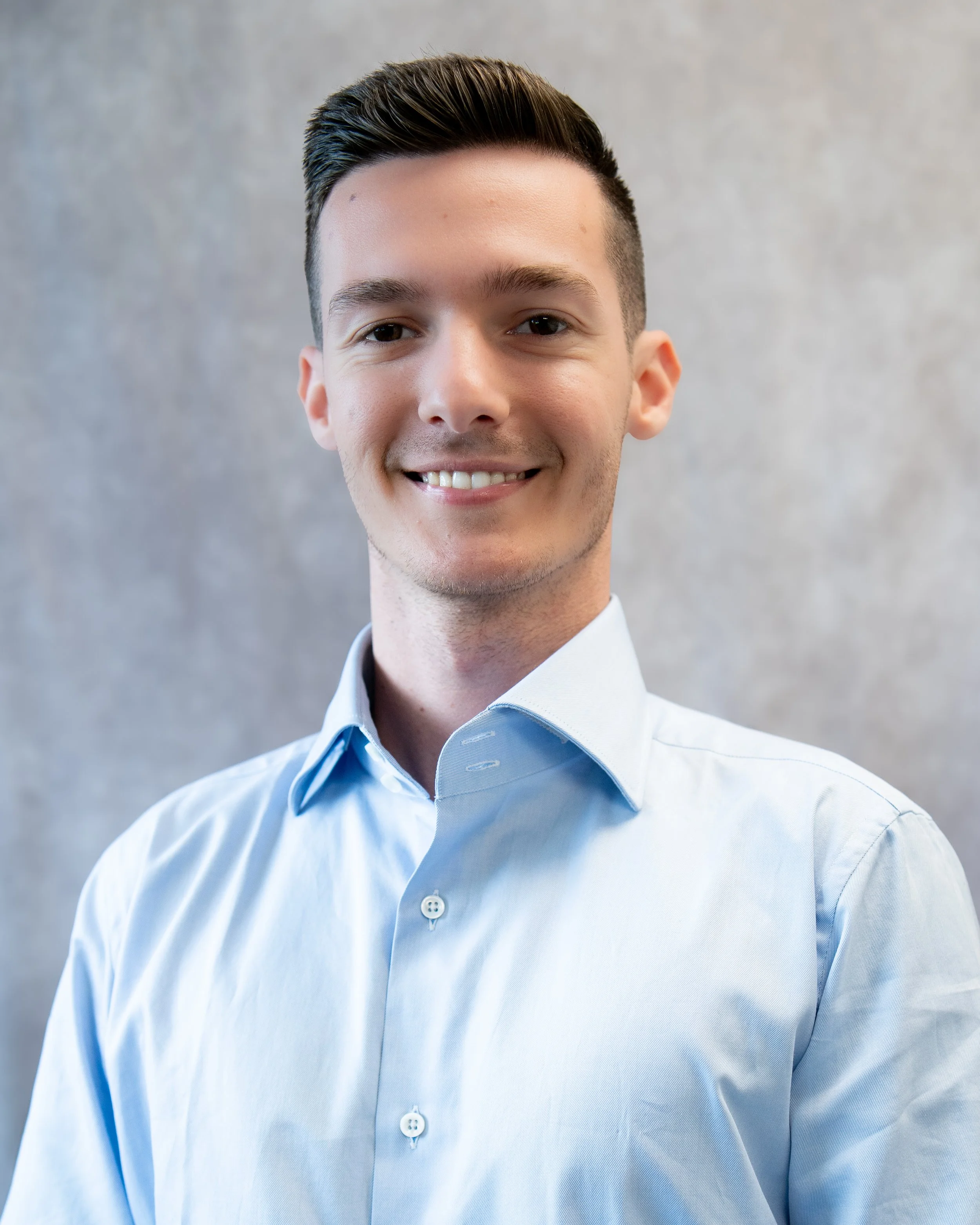 A young man with dark, slicked-back hair wearing a light blue button-up shirt, smiling at the camera, against a neutral gray background.